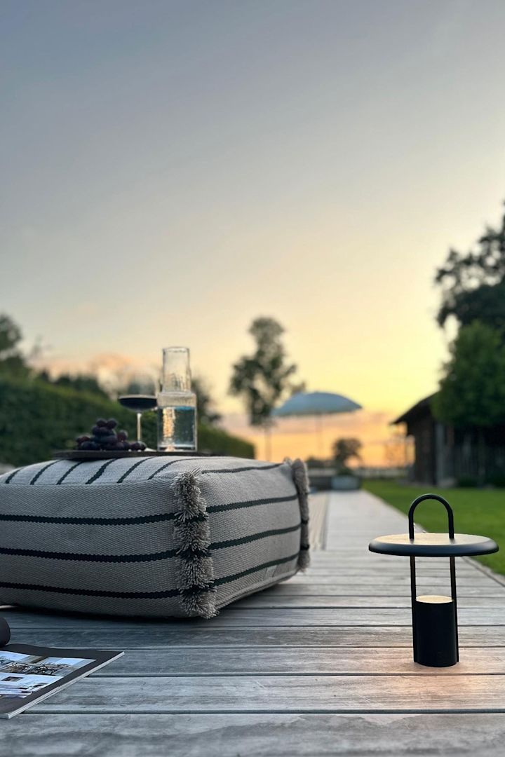 Een gezellig terras met een houten vloer, waarop het Lina zitkussen van OYOY ligt, naast een zwarte Pier tafellamp van Stelton.