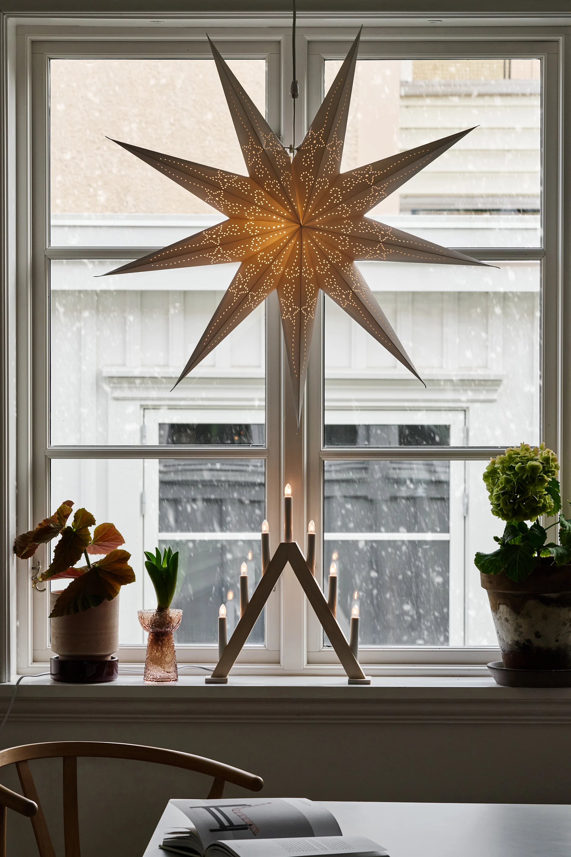 Een witte geplooide hanglamp hangt in een venster, geflankeerd door vitrages, boven een radiator en een kom met een bol.