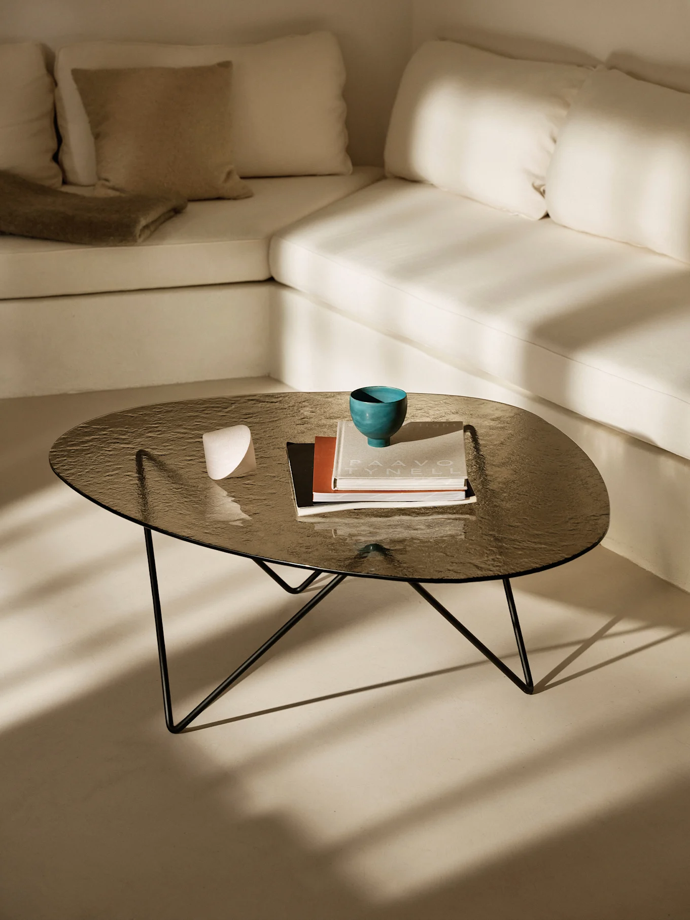 Donkere glazen salontafel met een teal kom, boeken en een wit object. Lichte L-vormige bank en zonlichtschaduwen.