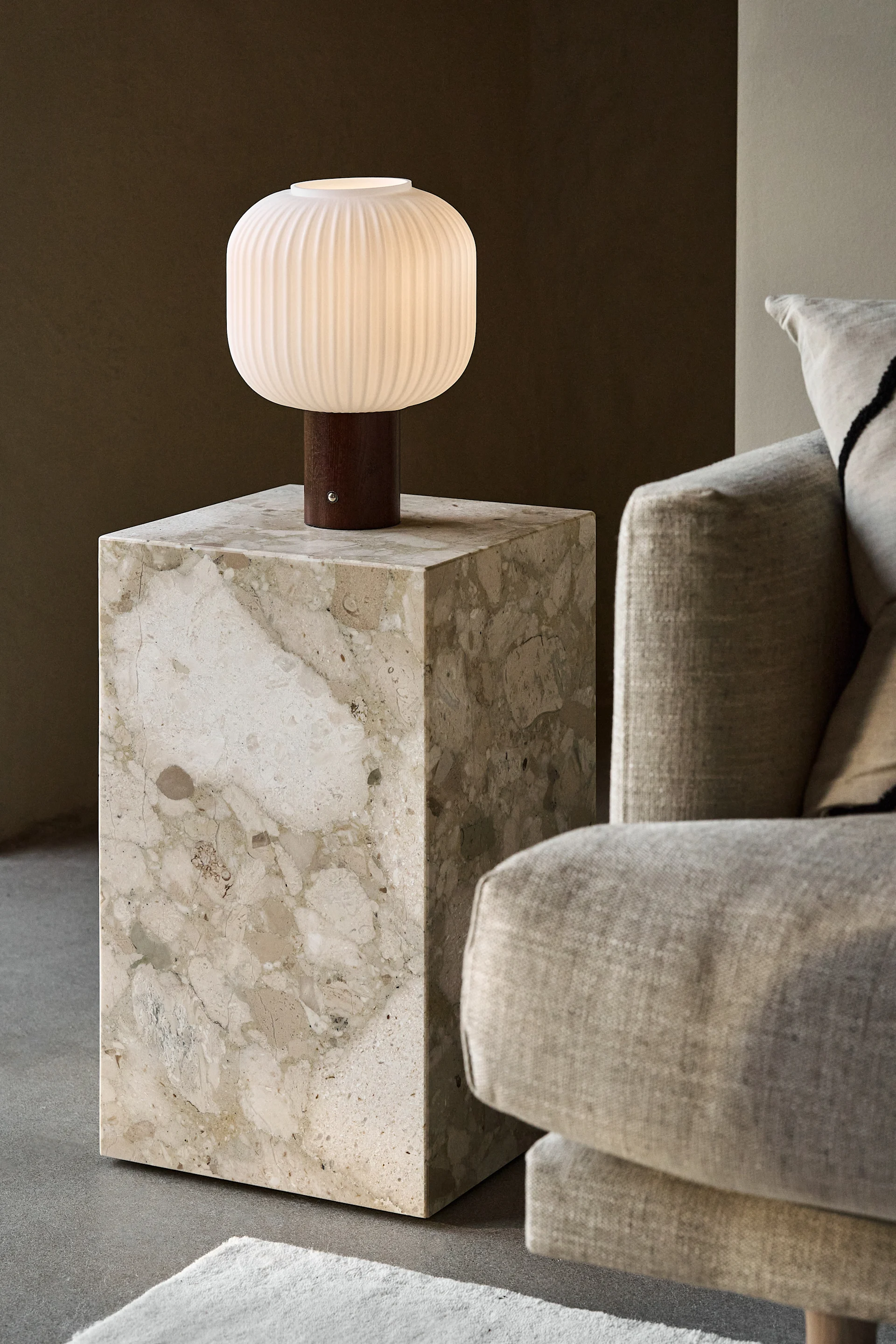 Een verlichte, geribbelde, witte tafellamp met houten voet op een beige marmeren bijzettafel naast een lichtgekleurde bank.