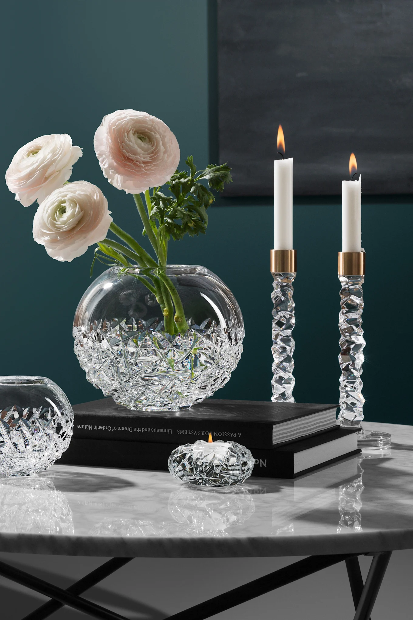 Elegante kristallen vaas met roze ranonkels, twee brandende kristallen kandelaars en een theelichthouder op een marmeren tafel.