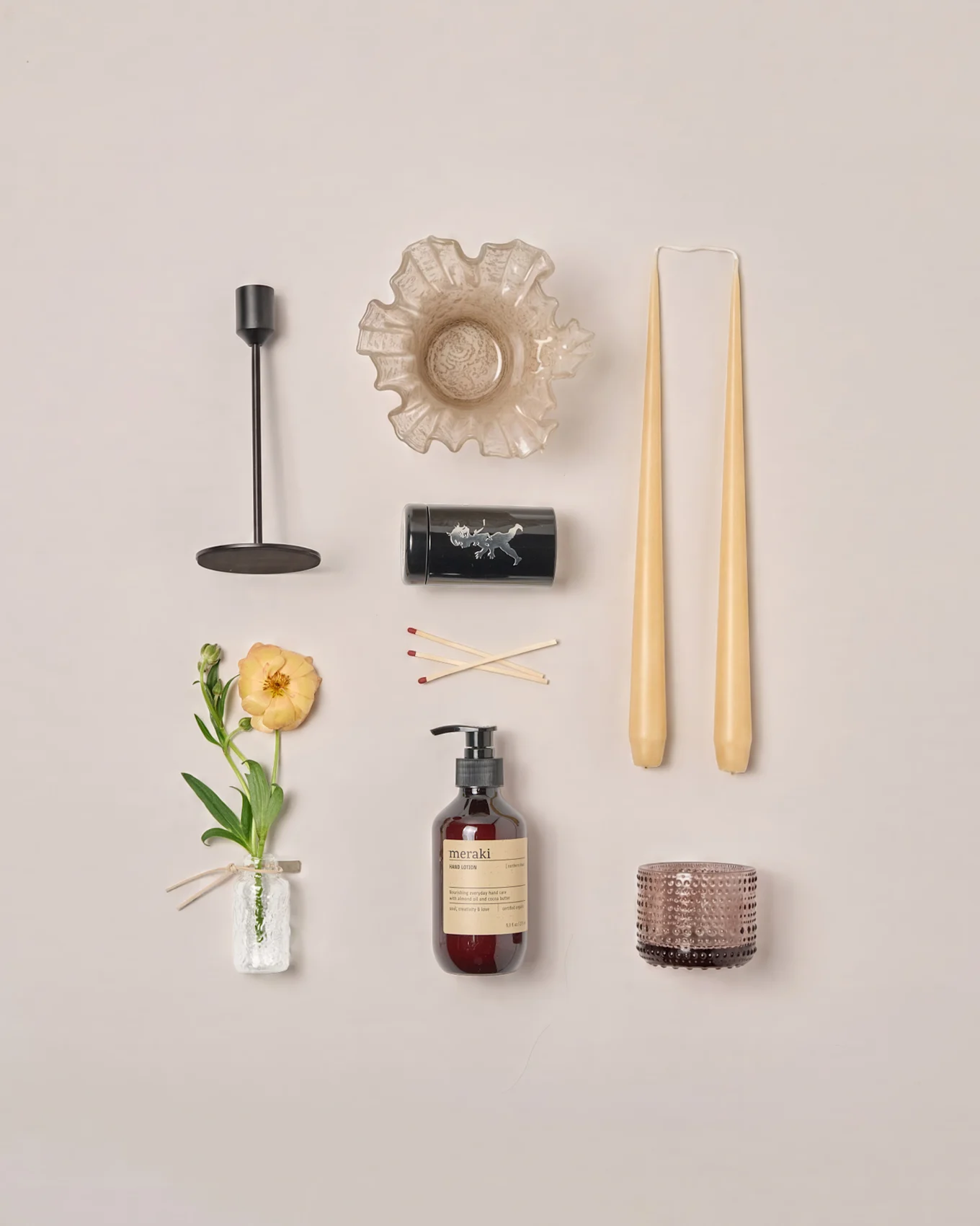 Een flat lay van diverse woondecoratieartikelen: kandelaar, gegolfde kom, taps toelopende kaarsen, lucifers, lotion, theelichthouder en een ranonkelbloem.
