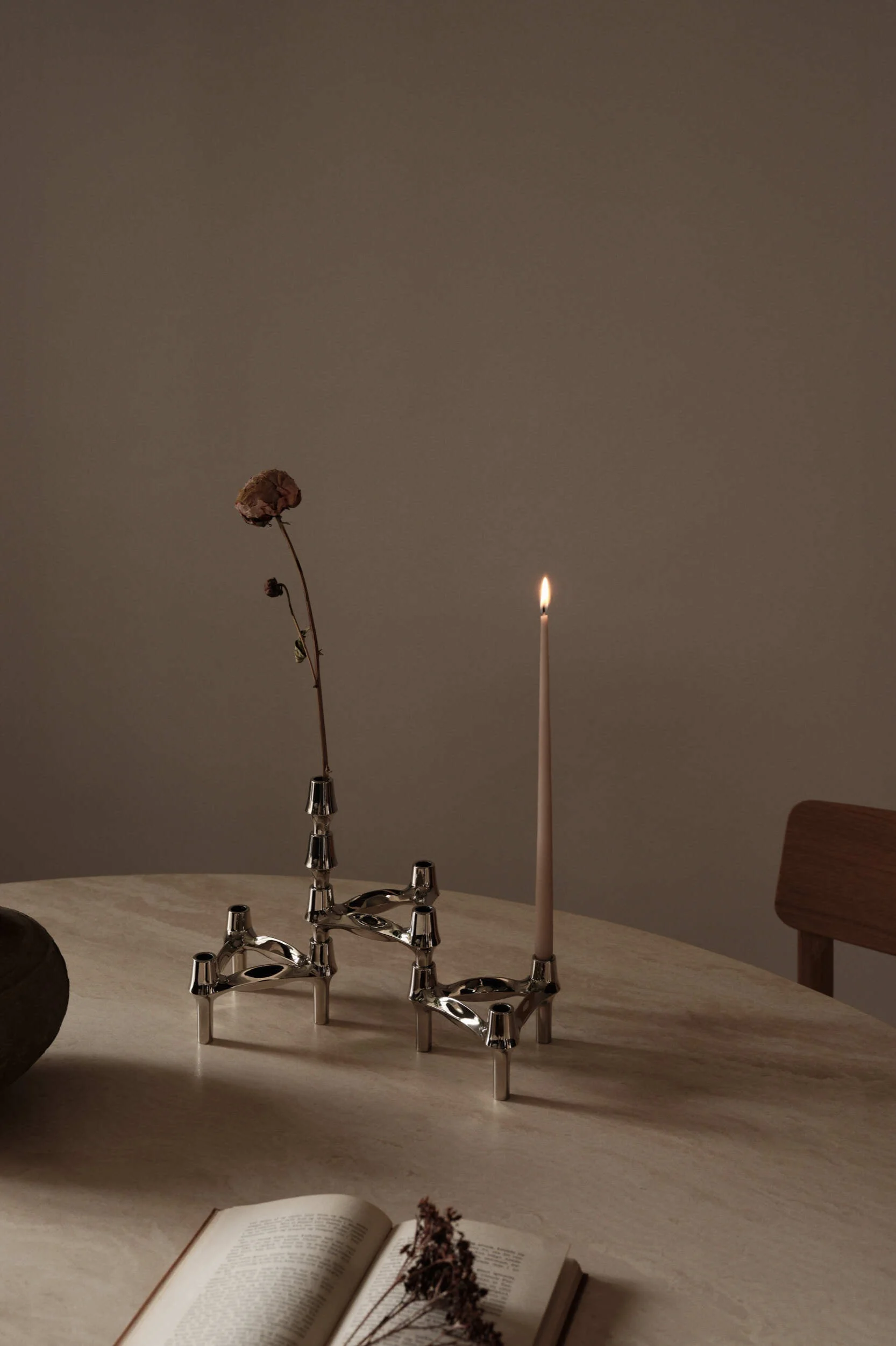 Twee zilverkleurige modulaire kandelaars op een ronde tafel, één met een brandende kaars, de andere met een gedroogde bloem. Een open boek is zichtbaar op de voorgrond.