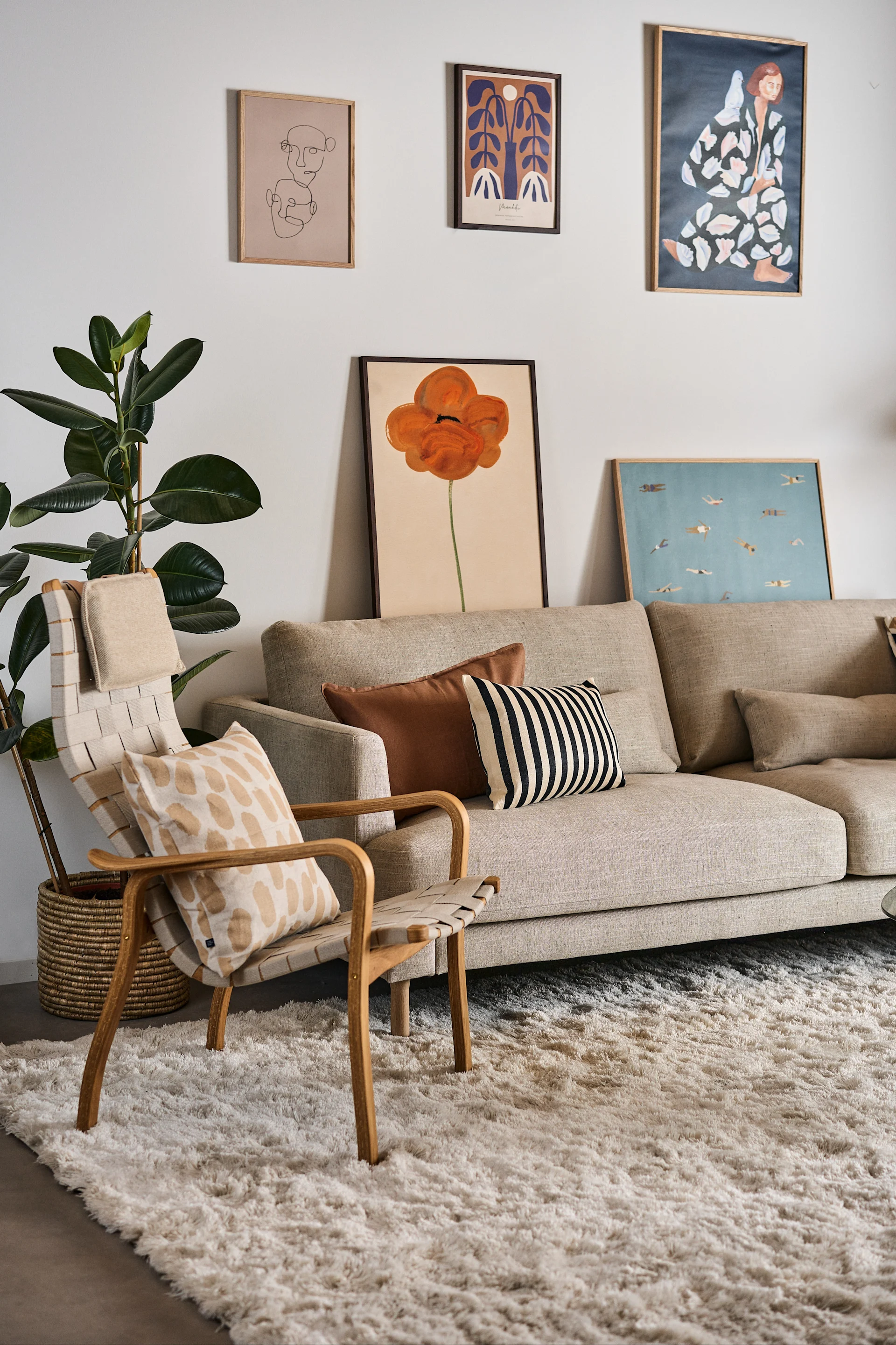 Moderne woonkamer met een beige bank, geweven fauteuil, grote groene plant, crèmekleurig vloerkleed en diverse ingelijste kunstprints.
