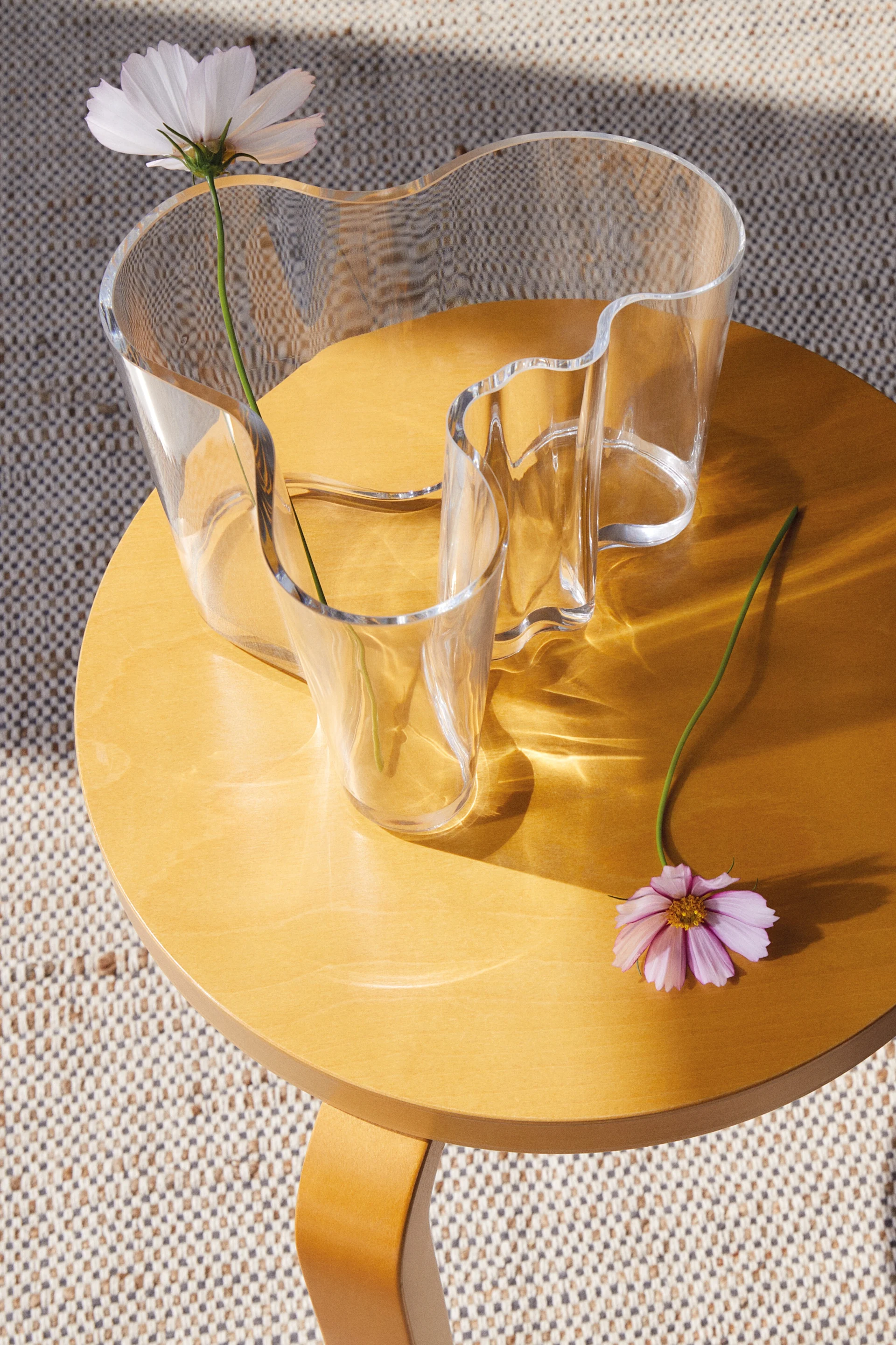 Een heldere, golvende glazen vaas met een witte bloem op een ronde houten tafel, met een roze bloem ernaast. Zonlicht werpt patronen.