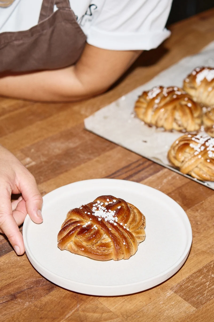 Hand plaatst een met suiker bestrooide, gevlochten kaneelbroodje op een bord; meer broodjes op een bakplaat.