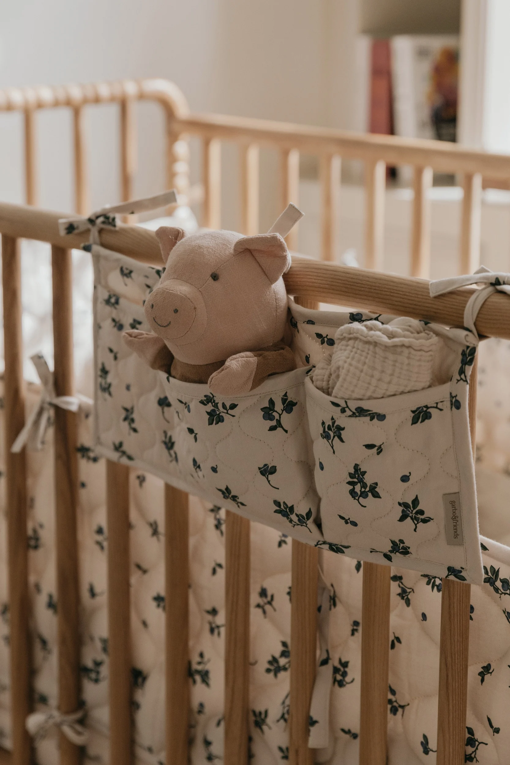 Een roze pluchen varken en een wit mousseline doekje in een crème-blauwe bloemen organizer aan een houten babybed.