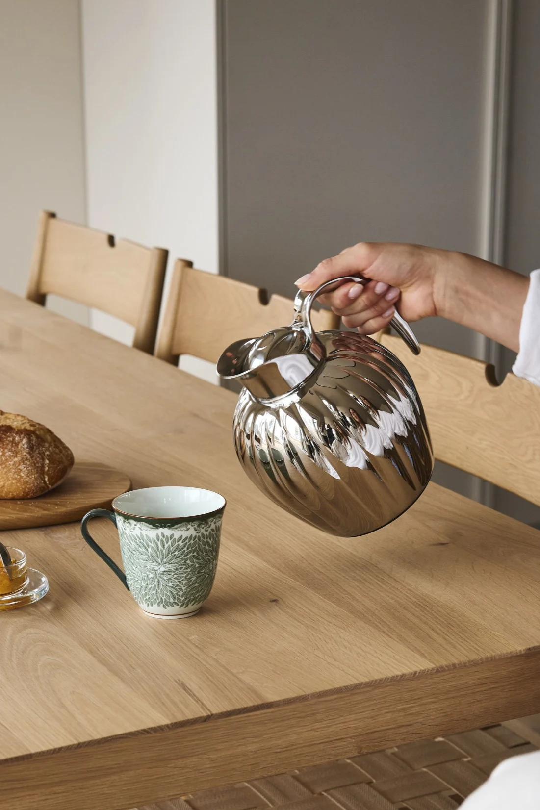 Een hand schenkt uit een zilveren geribbelde thermobeker in een groene mok op een houten tafel met brood en jam.