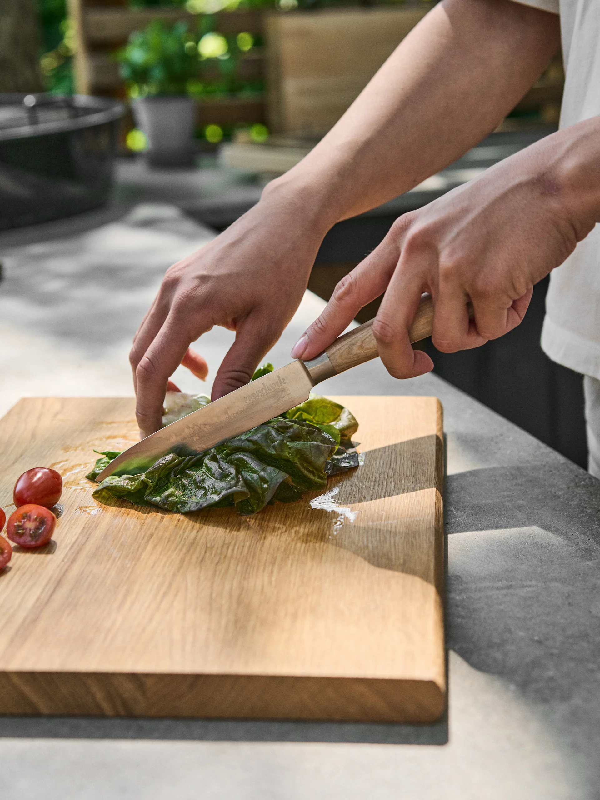 Handen snijden een groen blad met een mes op een houten snijplank, naast cherrytomaten.