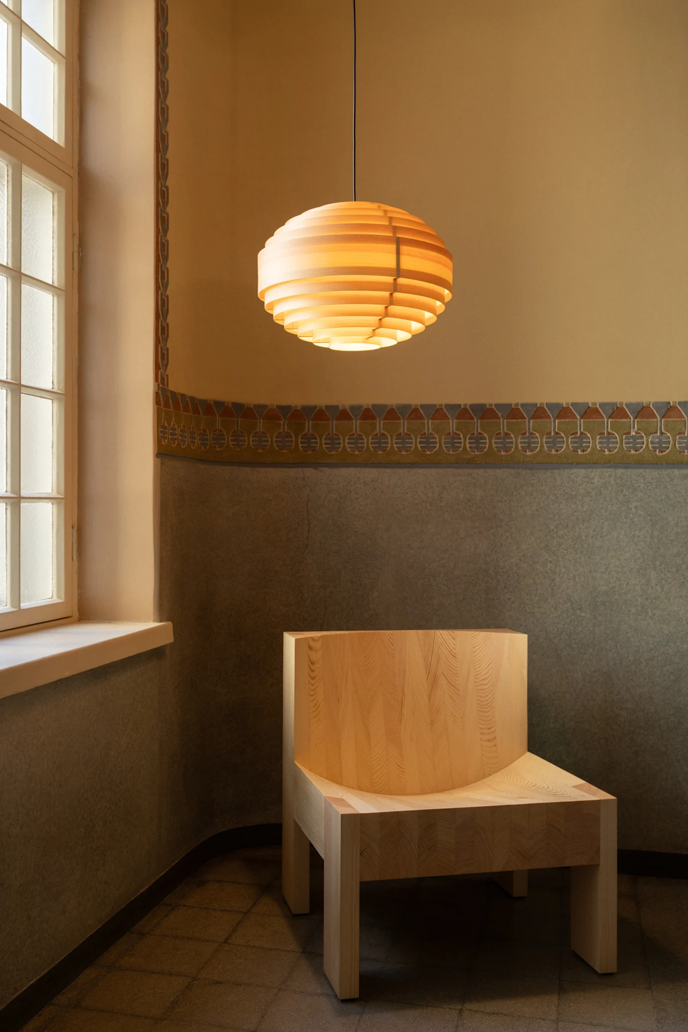 Houten lamellen hanglamp boven lichte houten stoel in een kamer met gedessineerde muren en een raam.