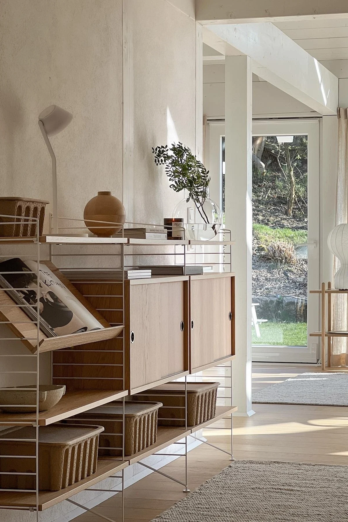 Lichte, moderne kamer met rekkensysteem van licht hout en witte frames, gevuld met vaas, boeken, planten en manden, met tuinzicht.