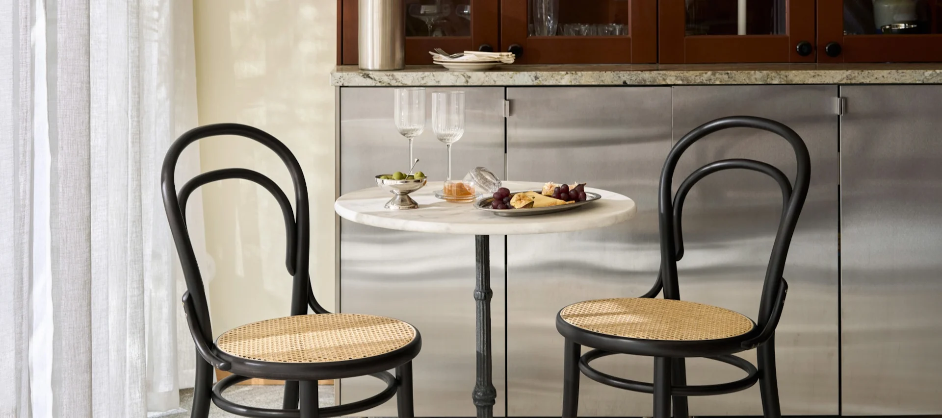 Twee zwarte bistrostelen met rotan zittingen aan een ronde marmeren tafel met wijnglazen, olijven, druiven en kaas.