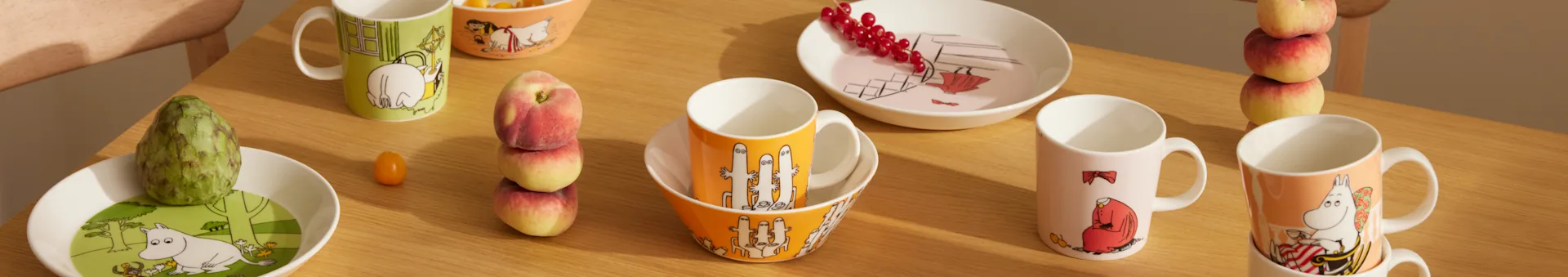 Moomin servies in diverse kleuren en ontwerpen, met mokken, kommen en borden, samen met vers fruit op een houten tafel.