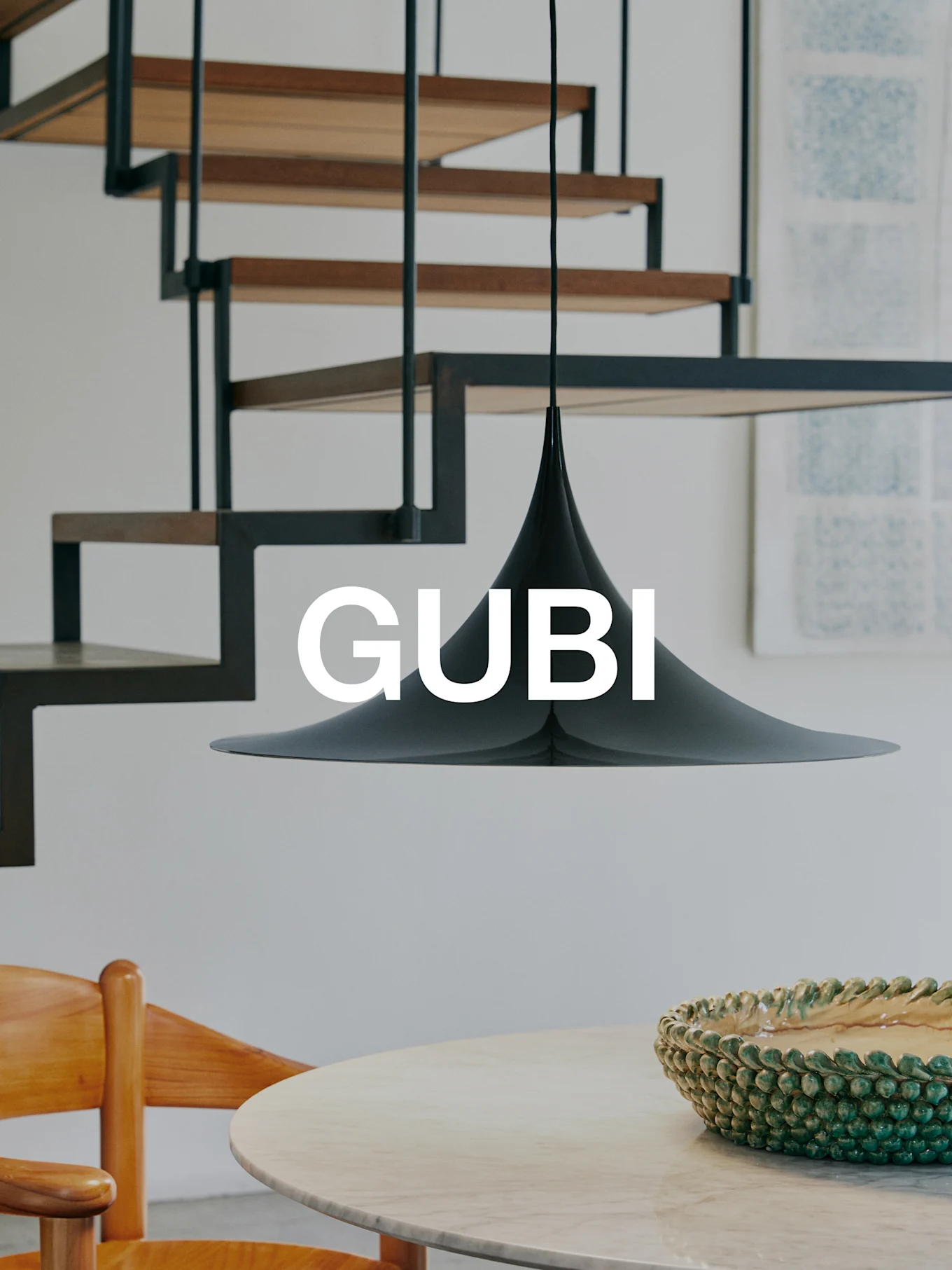 Een zwarte conische hanglamp boven een marmeren tafel met een groene kom en een houten stoel. Een moderne trap op de achtergrond. Tekst 'GUBI'.