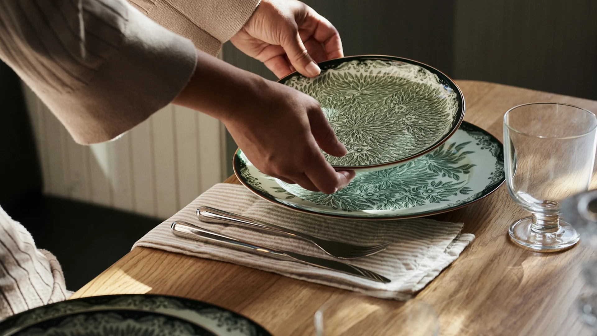 Handen plaatsen een groen-wit patroonbord op een houten tafel met bestek en een glas.