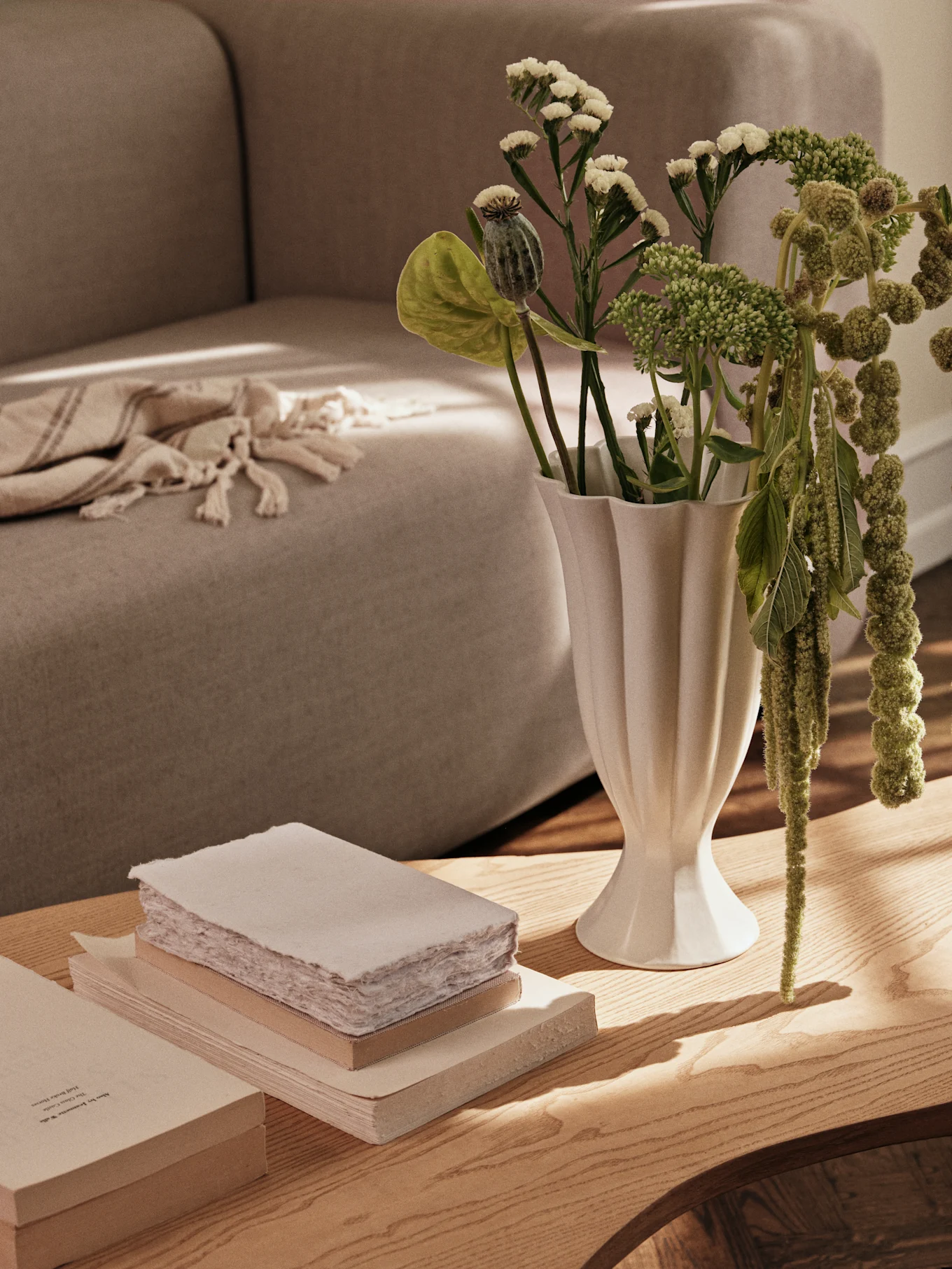 Een witte geribbelde vaas met groene planten en een stapel boeken op een houten salontafel, met een bank op de achtergrond.