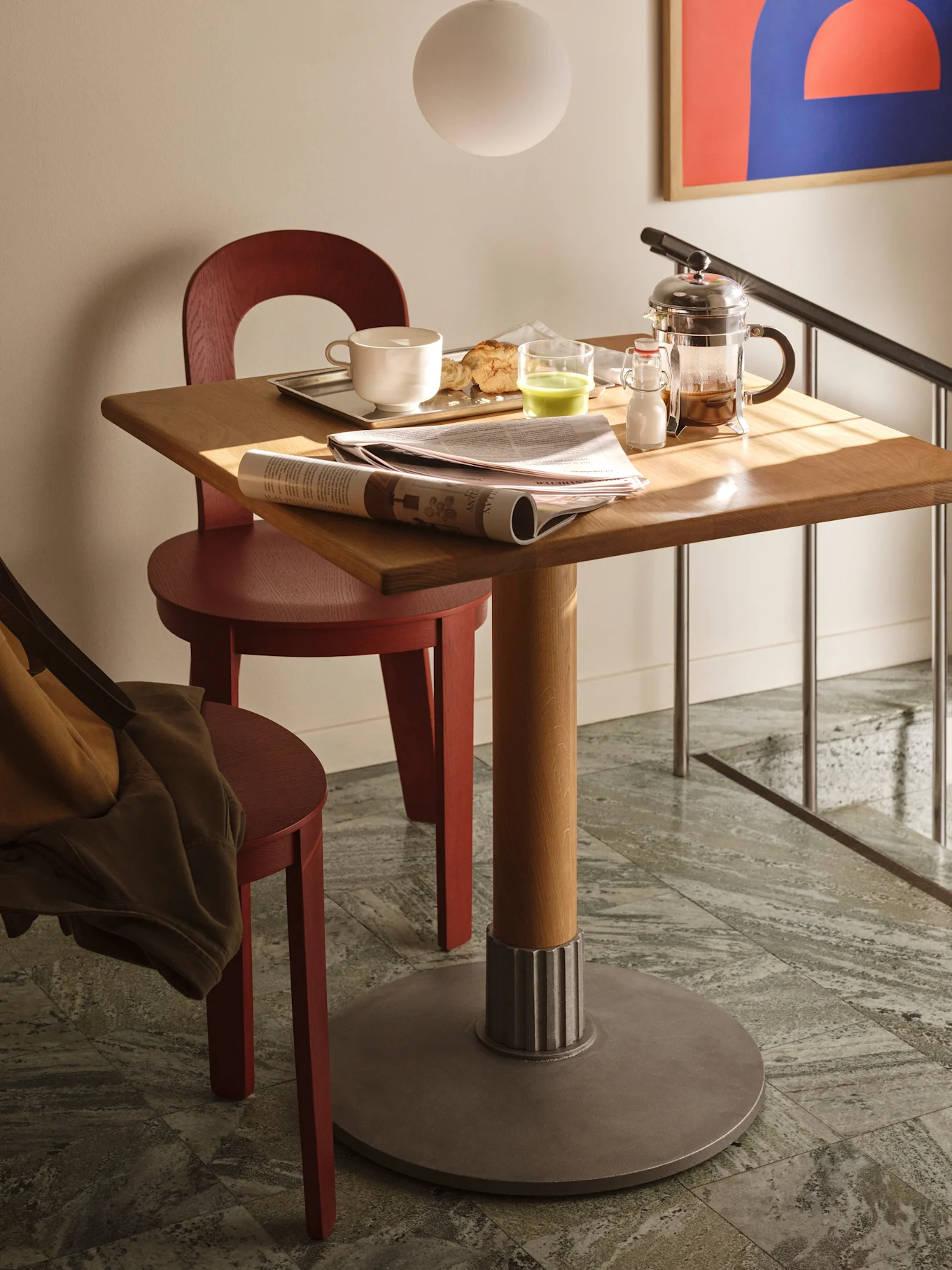 Een vierkante houten tafel met ontbijt, een krant en een cafetière, geflankeerd door twee rode houten stoelen op een tegelvloer.