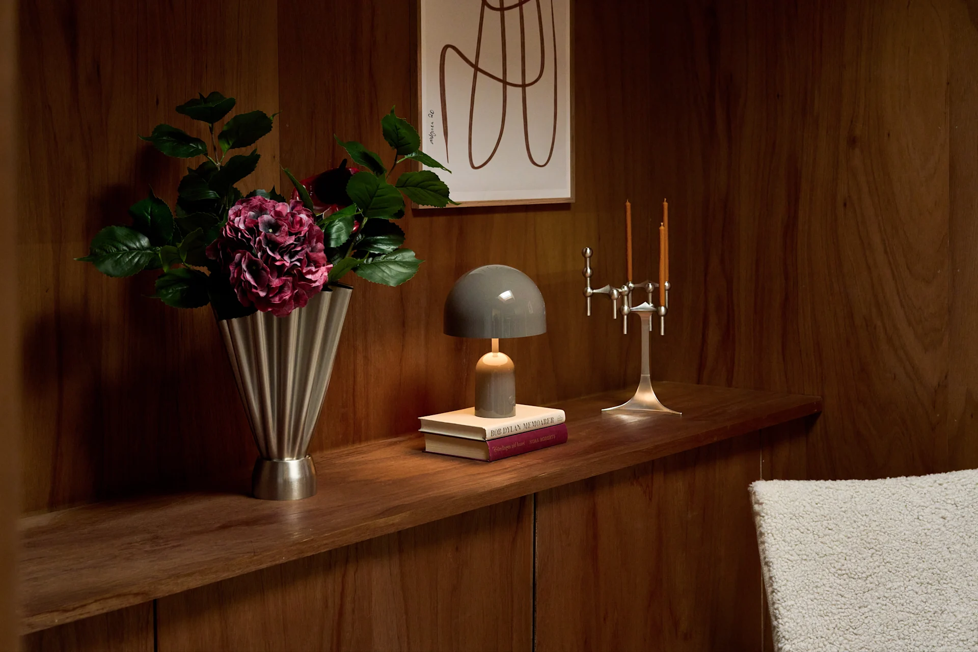 Houten plank: zilveren vaas met paarse bloemen, grijze paddenstoellamp op boeken, zilveren kandelaar met twee brandende kaarsen.