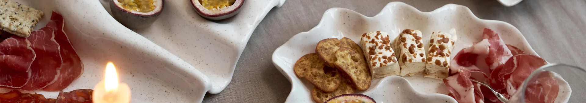 Een voorgerechtenplank op twee schelpvormige schalen met vleeswaren, crackers, kaas en passievrucht, met een brandende kaars.