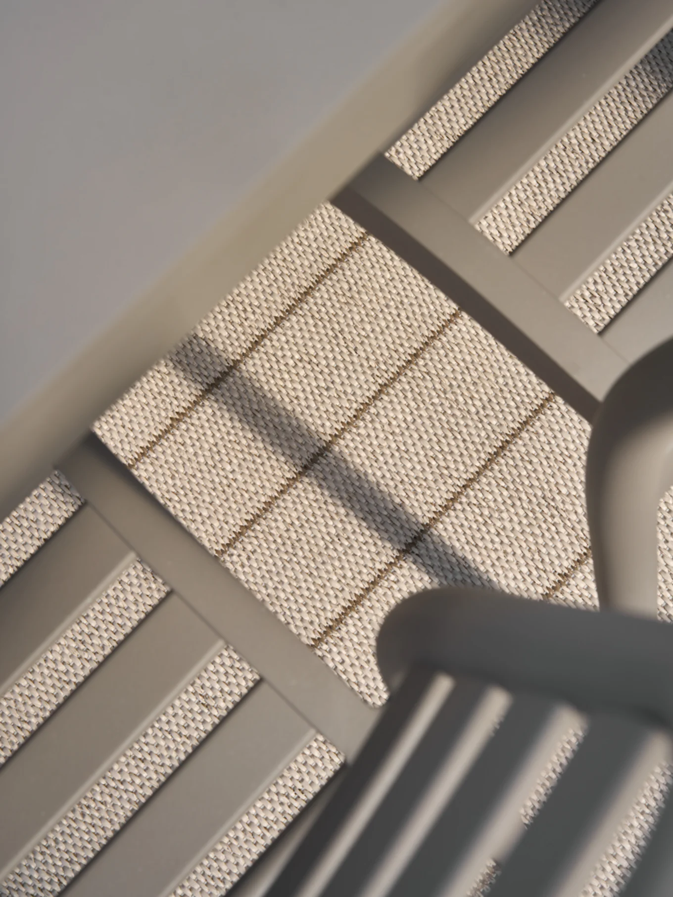 Close-up, schuine weergave van lichtbeige geweven materiaal met subtiele horizontale en diagonale bruine strepen, omlijst door gladde grijze staven.