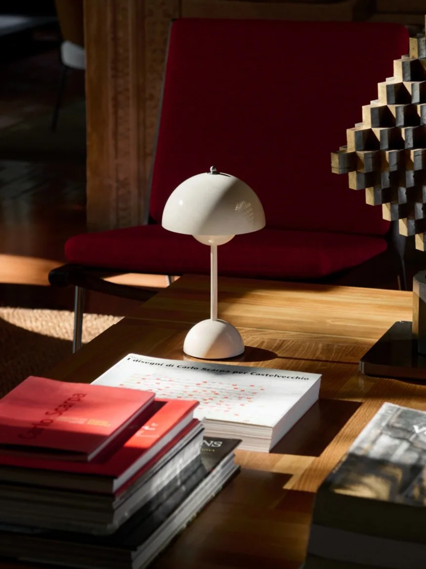 Een witte paddestoelvormige tafellamp, boeken en een geometrische sculptuur op een houten salontafel, met een rode fauteuil erachter.