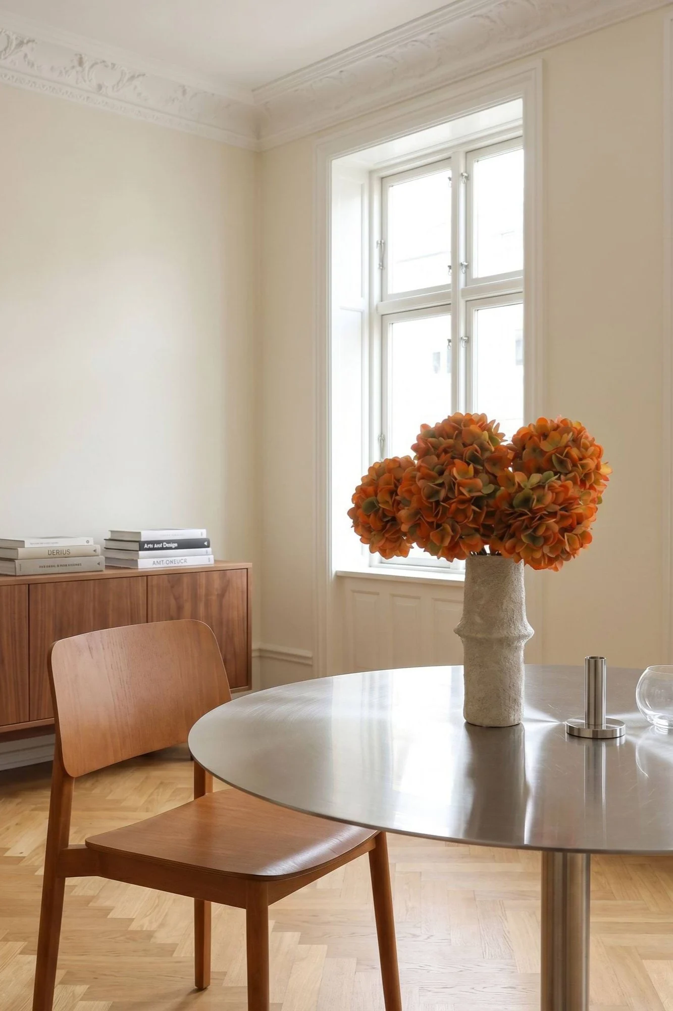 Een lichte eetkamer met een ronde metalen tafel, een houten stoel en een vaas met oranje bloemen.