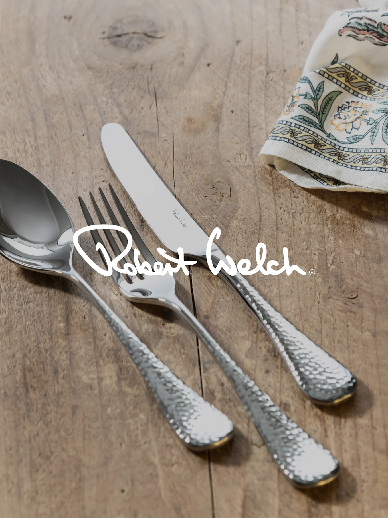 Robert Welch logo over zilveren gehamerd bestek (lepel, vork, mes) op hout met een gedessineerde doek.