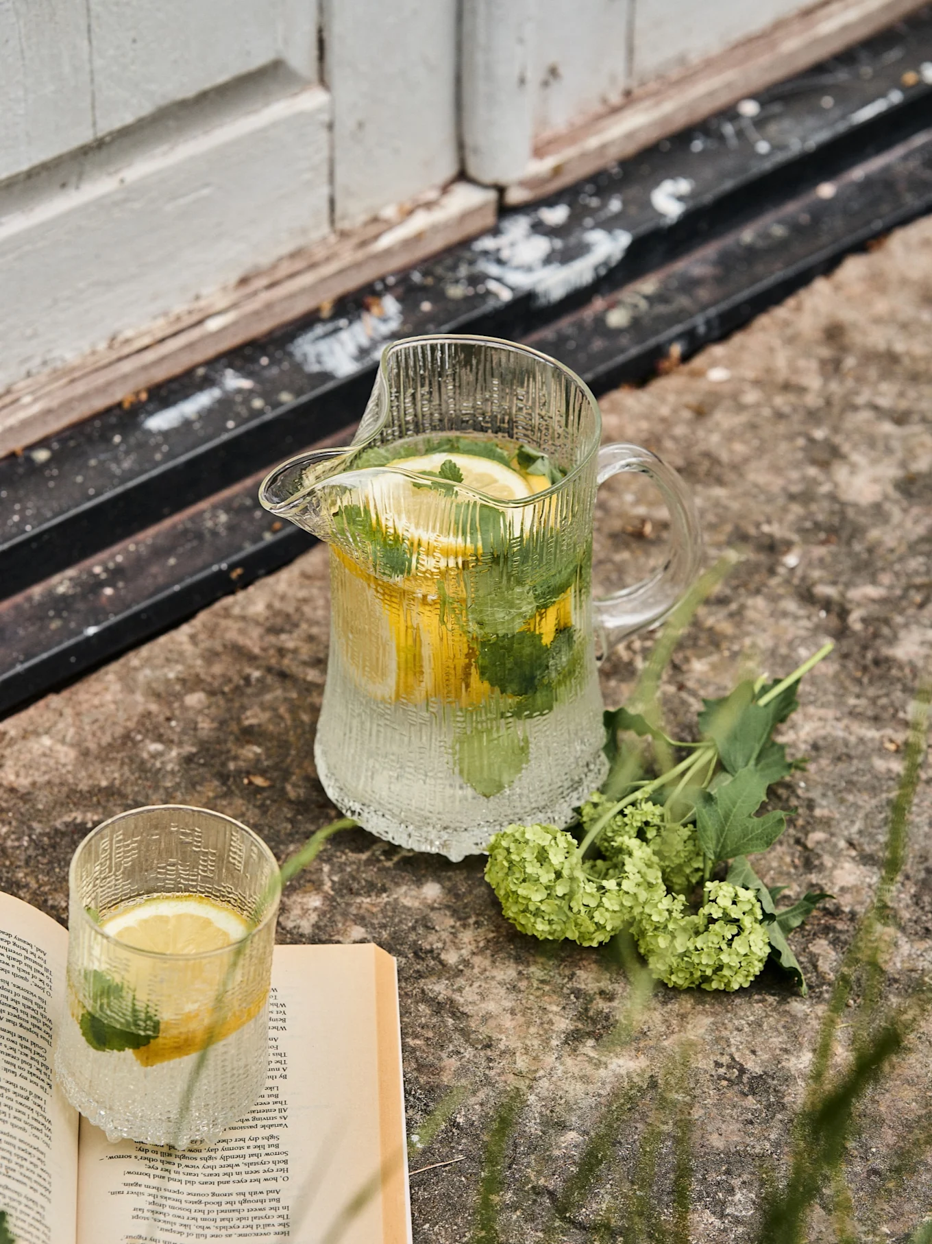 Geribbelde glazen kan en bijpassend glas met citroen- en muntwater, naast een open boek en groene bloemen op steen.