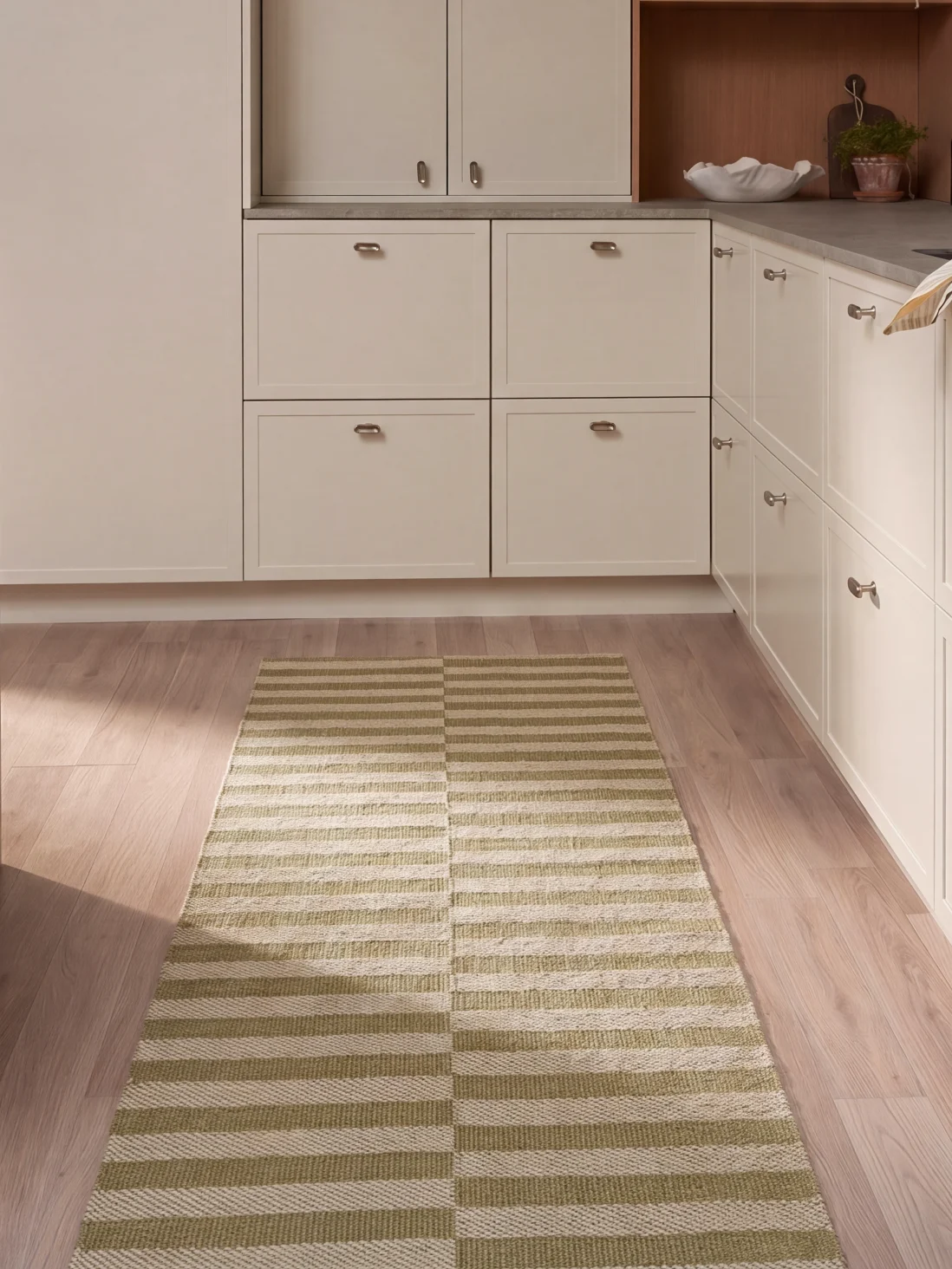 Een keuken met lichte kasten, houten vloer en een lang rechthoekig tapijt met groene en witte horizontale strepen.
