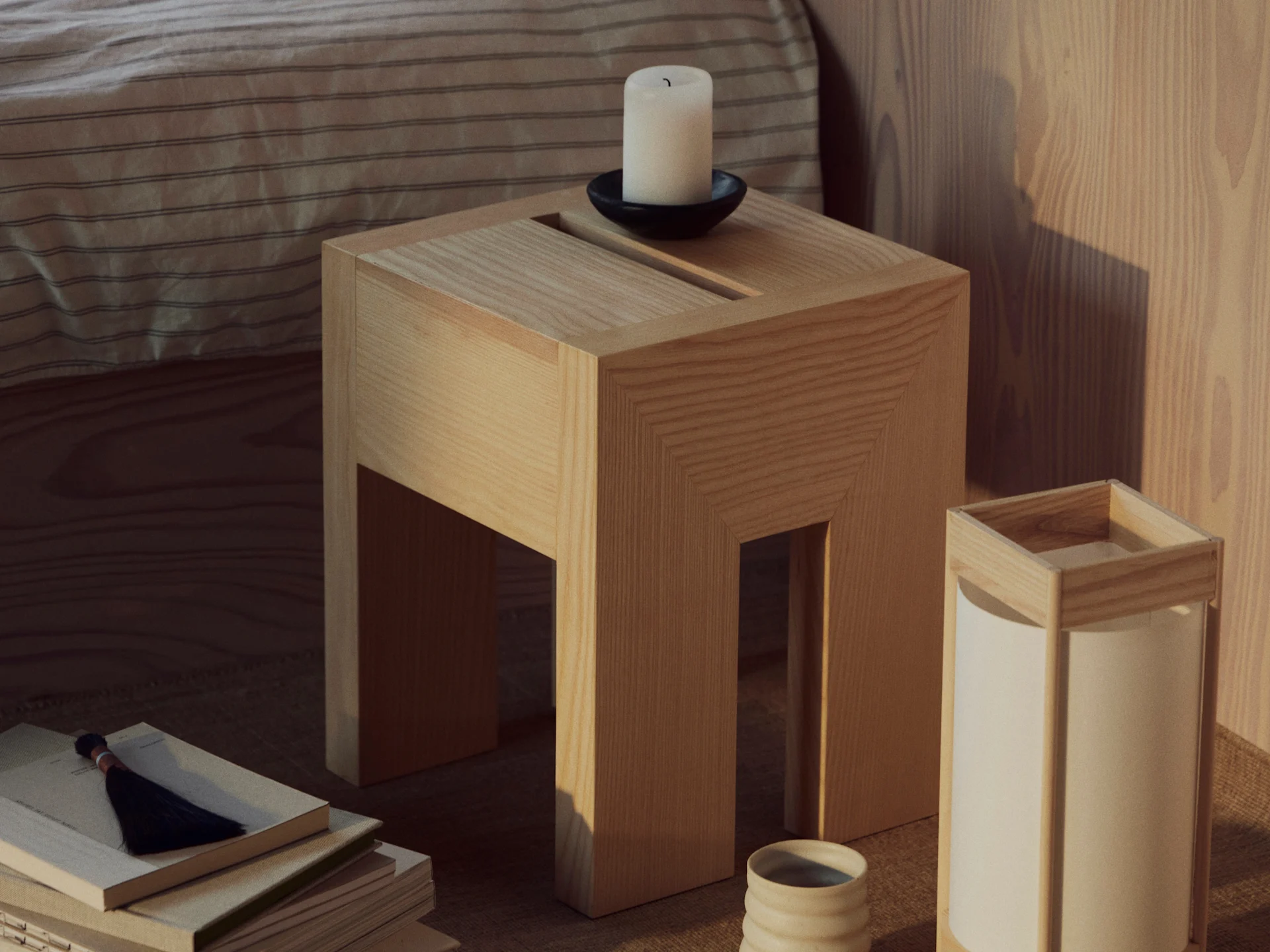 Een houten bijzettafel met een witte kaars in een zwarte houder, een stapel boeken met een kwast en een lamp met een keramische beker.