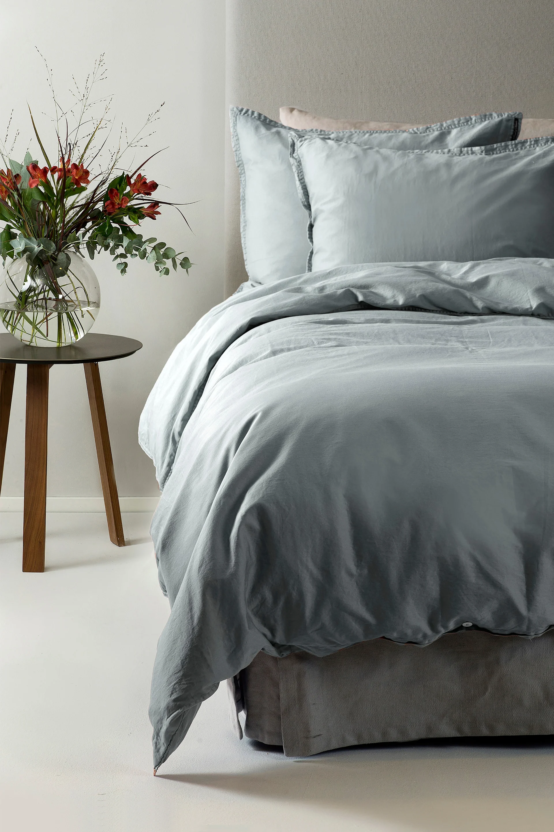 Een bed met lichtblauw beddengoed en twee kussens, naast een houten bijzettafel met een vaas rode bloemen.
