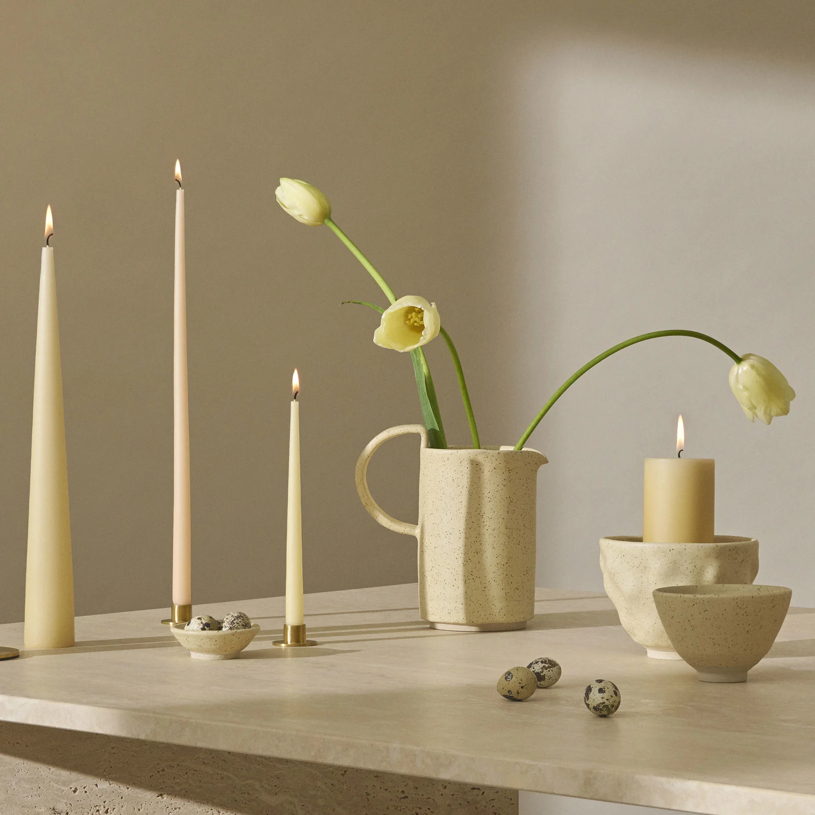 Een stilleven met brandende kaarsen, lichtgele tulpen in een gespikkelde kan en kwarteleitjes op een gestructureerd oppervlak.