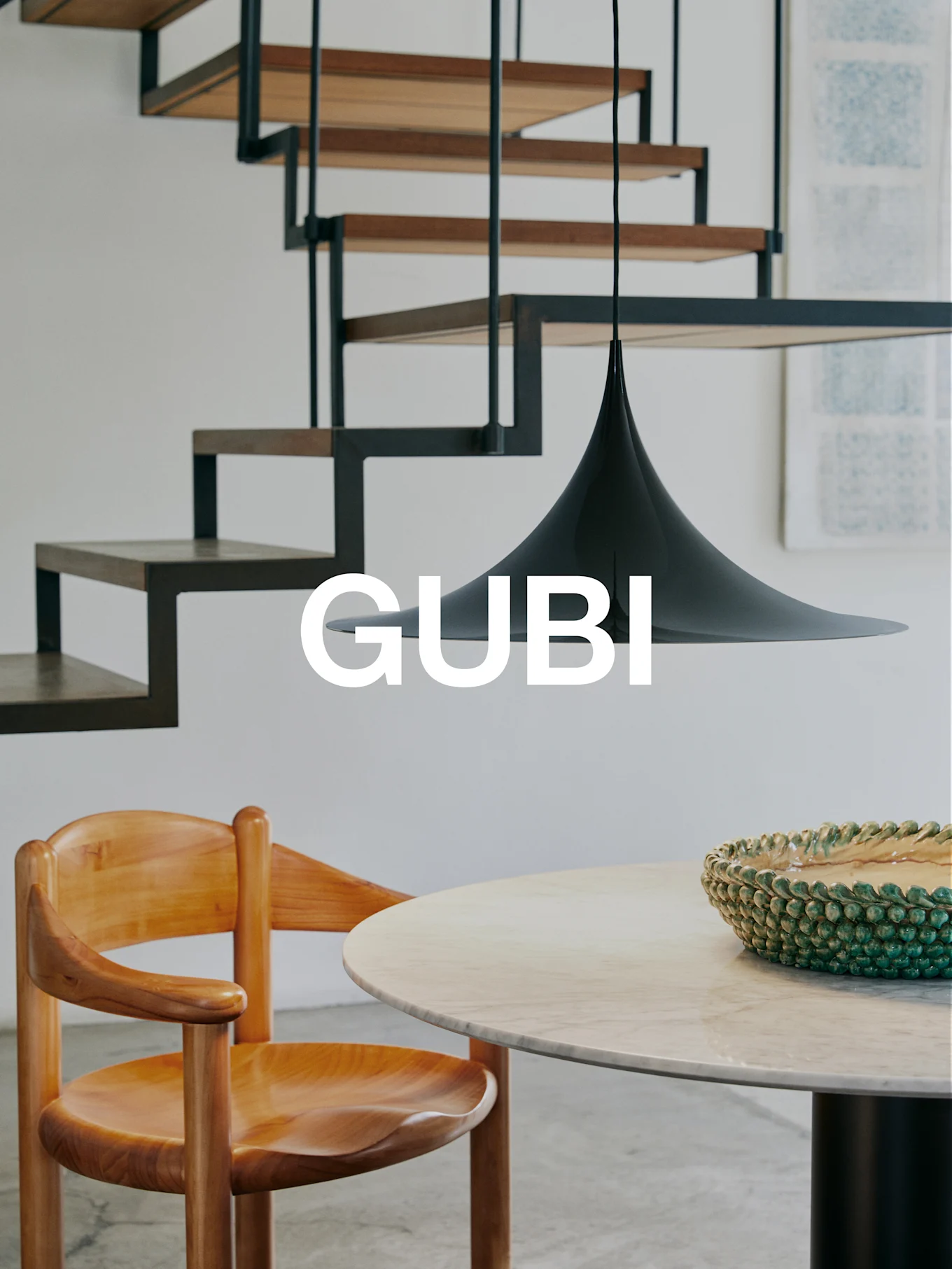GUBI-logo in een modern interieur met houten stoel, marmeren tafel, groene kom, zwarte hanglamp en moderne trap.