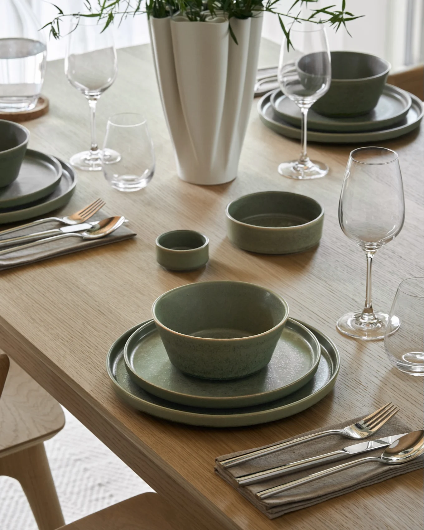 Eettafel gedekt met kommen romige soep, bijpassende borden, wijn- en waterglazen, bestek en een vaas met groen loof.