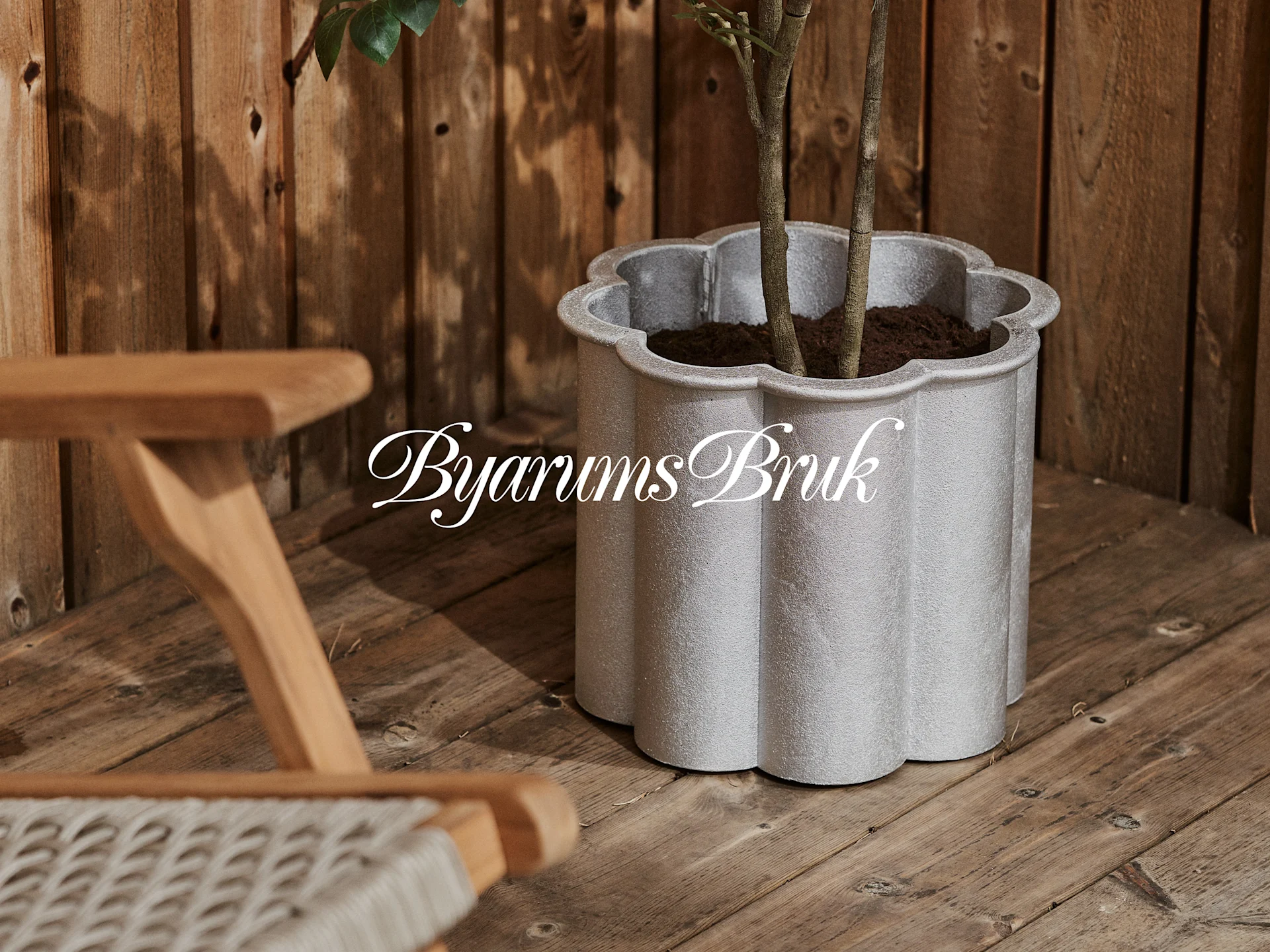 Een zilvergrijze, geschulpte plantenbak met een plant op een houten terras, naast een houten stoel. 'Byarums Bruk' logo.