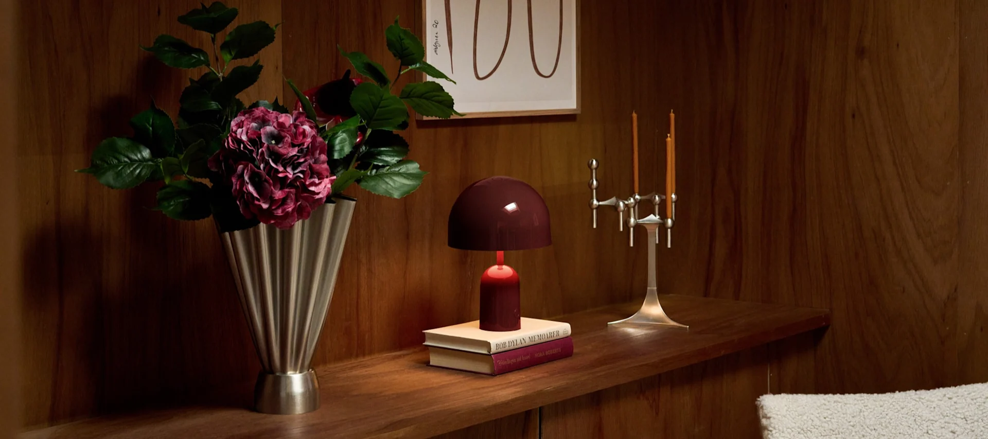 Houten plank: zilveren vaas met donkere hortensia's, bordeauxrode koepellamp op boeken, zilveren kandelaar met oranje kaarsen.