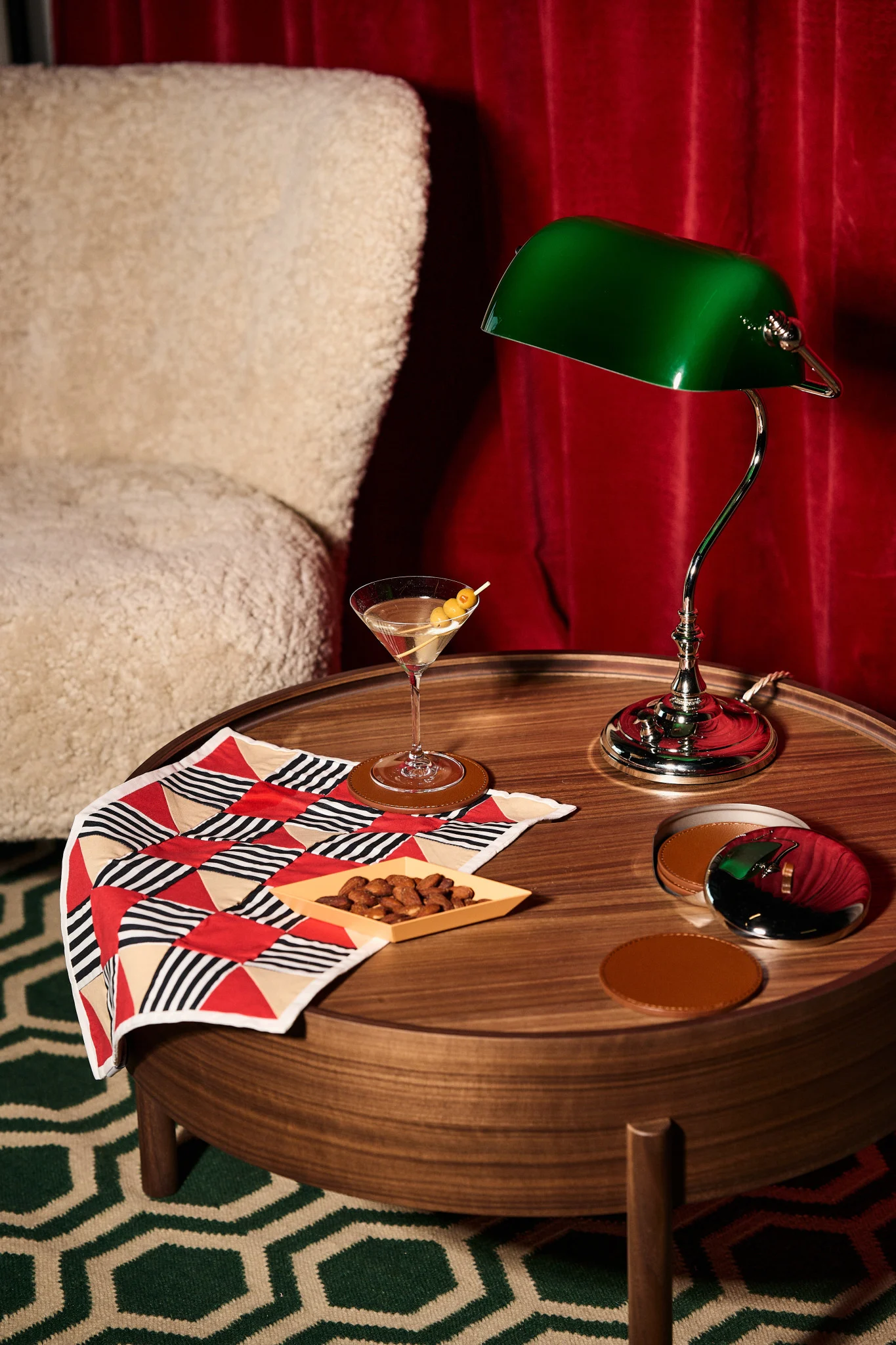 Martini, amandelen, groene bankierslamp op een houten tafel met geometrische doek, crème fauteuil en rode gordijnen.
