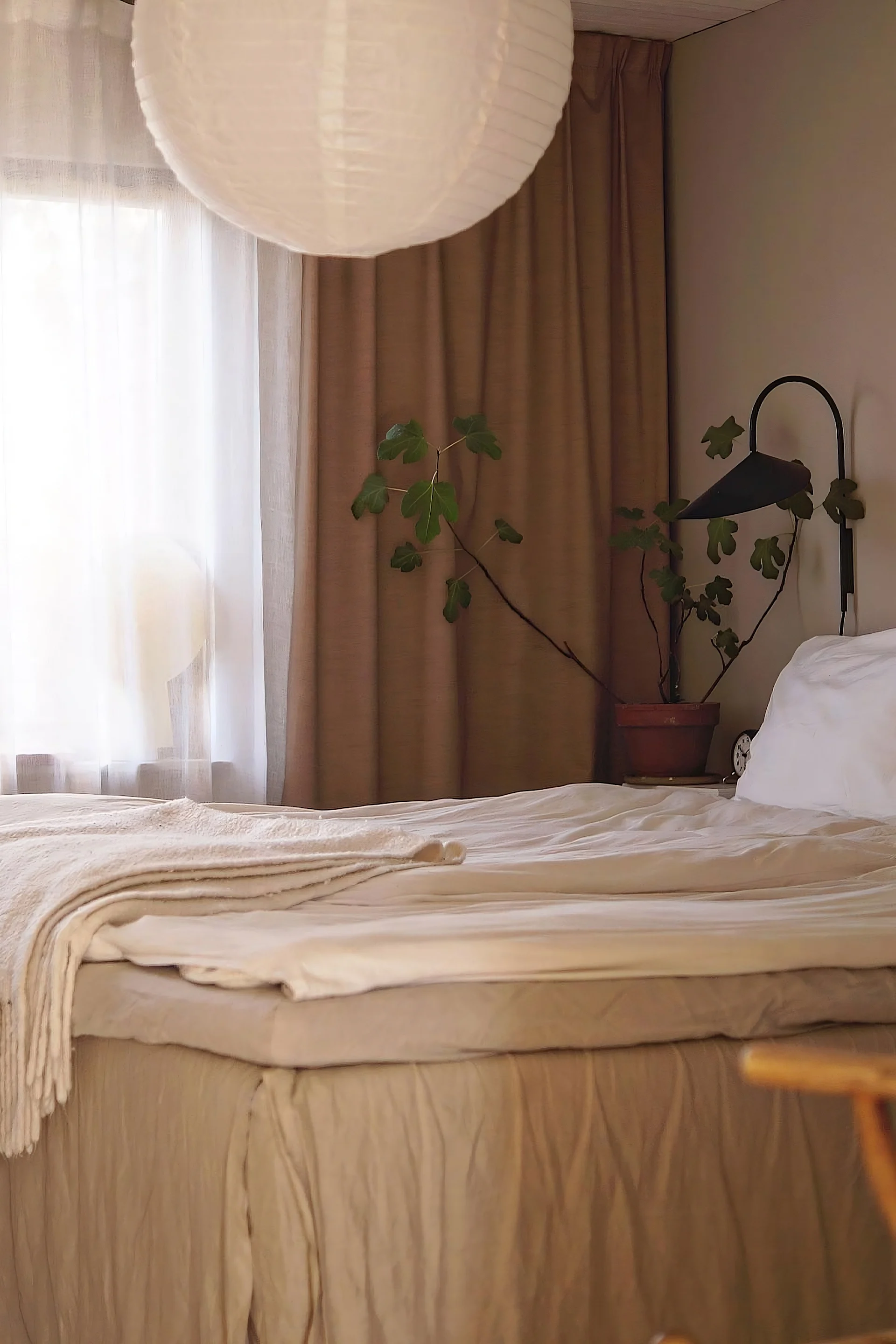 Een gezellige slaapkamer met een net opgemaakt bed in neutrale tinten, een grote papieren lantaarn, een groene potplant en een zwarte wandlamp.