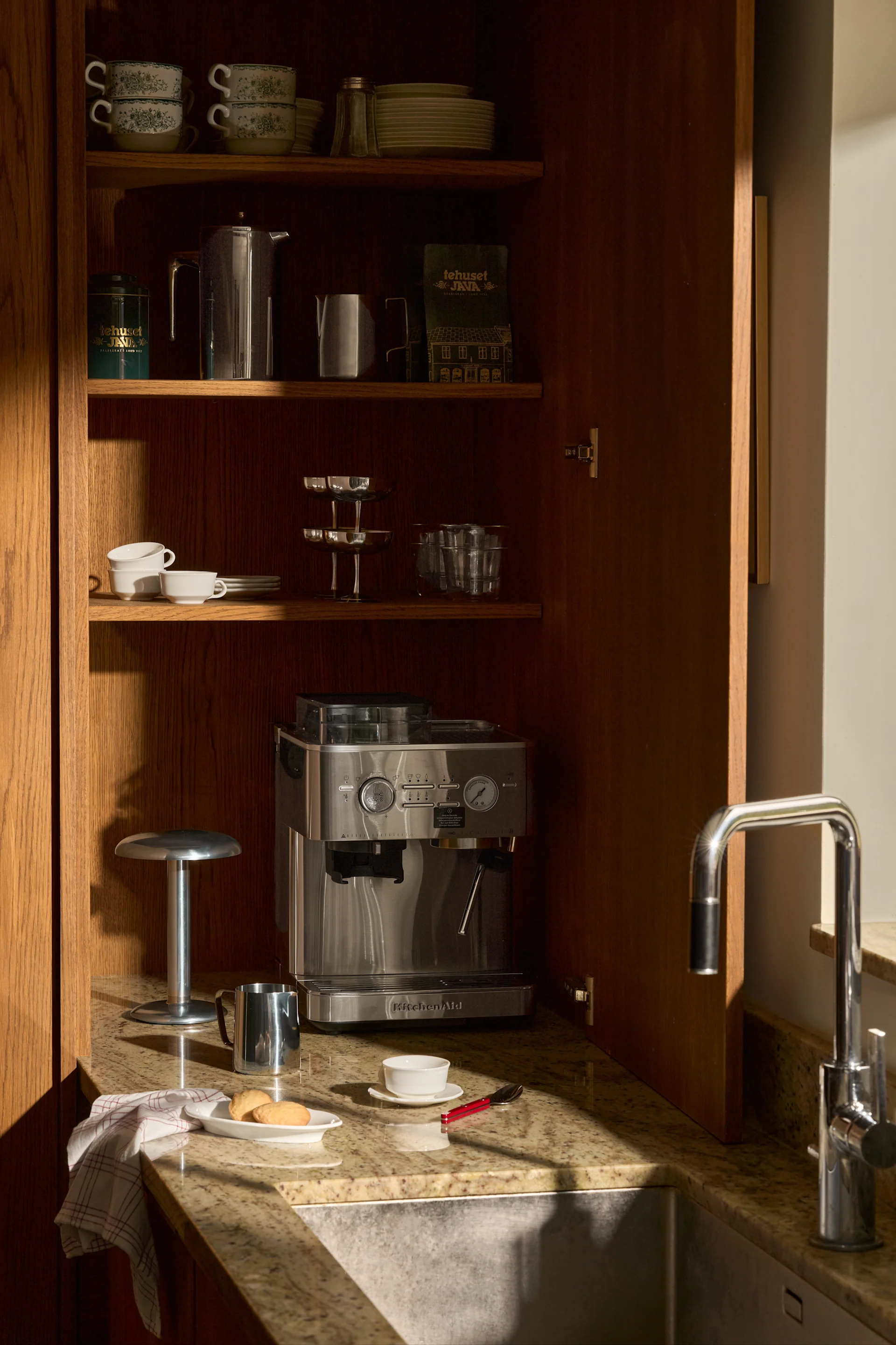 Open houten kast: KitchenAid espressomachine, gestapelde kopjes, borden, koffieartikelen en spoelbak op graniet.