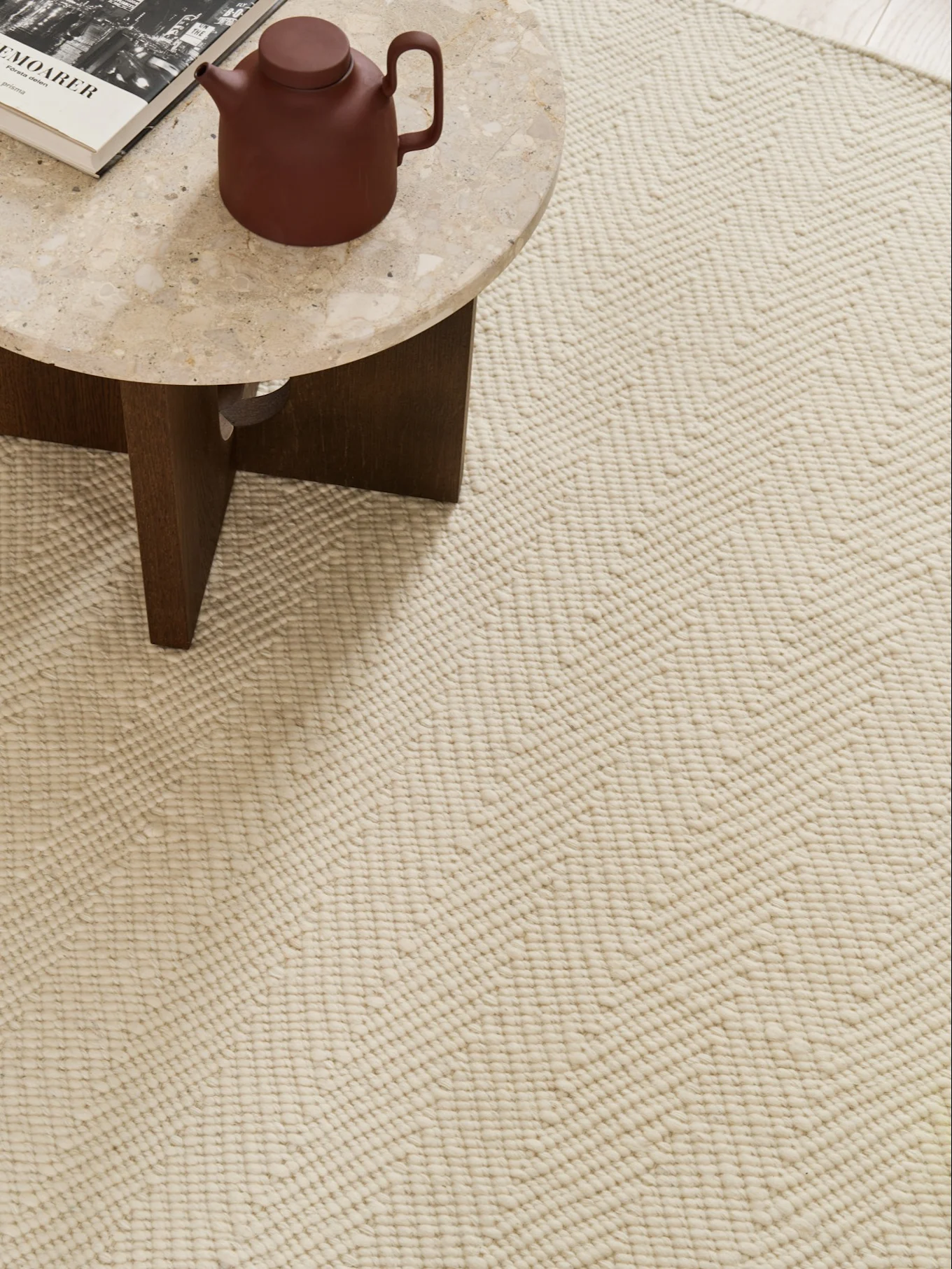 Close-up van een crèmekleurig tapijt met een getextureerd zigzagpatroon, met een marmeren tafel met een theepot en boek in de hoek.