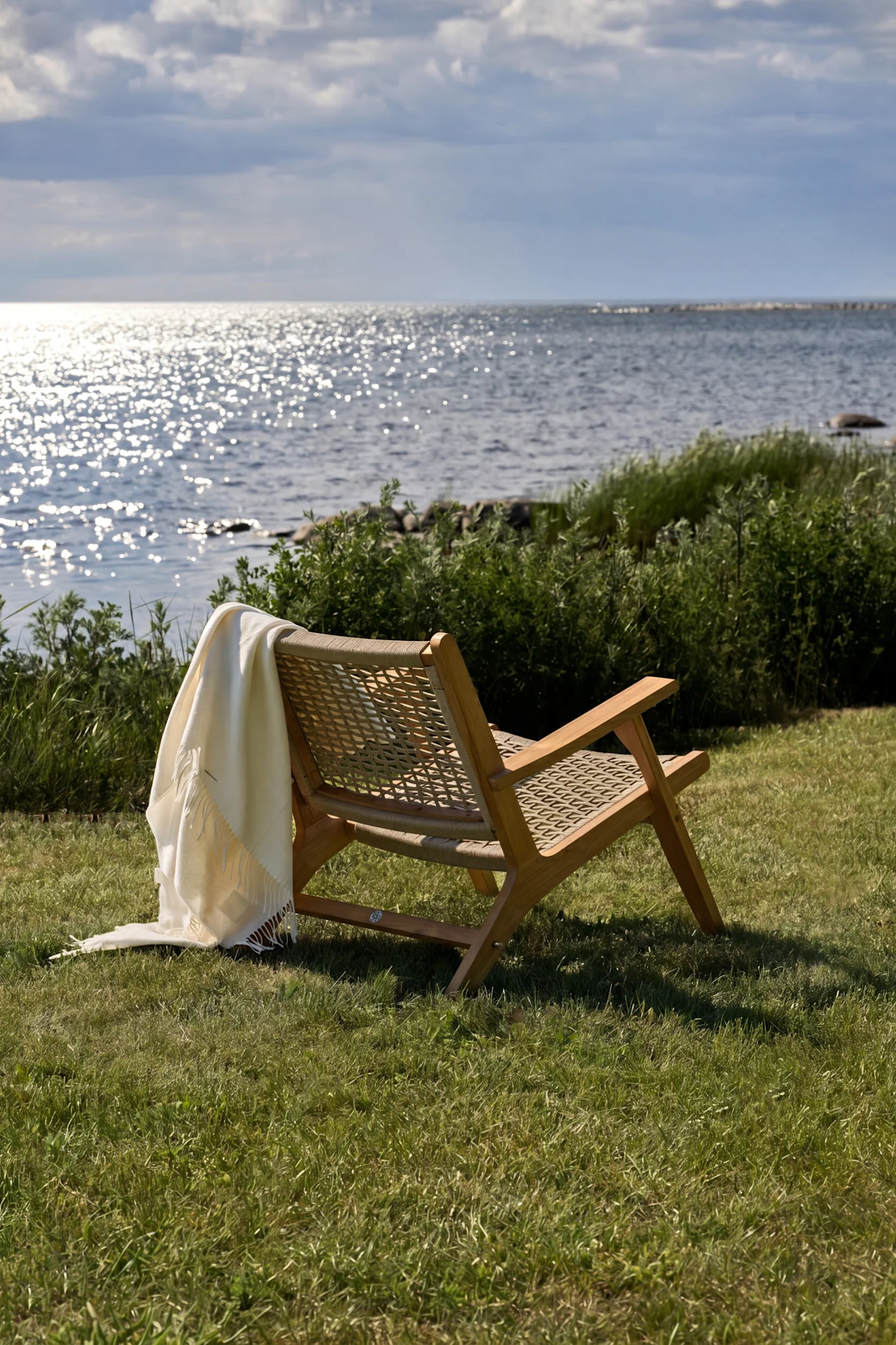 Houten loungestoel met een witte plaid op groen gras, gericht op sprankelend water onder een bewolkte lucht.