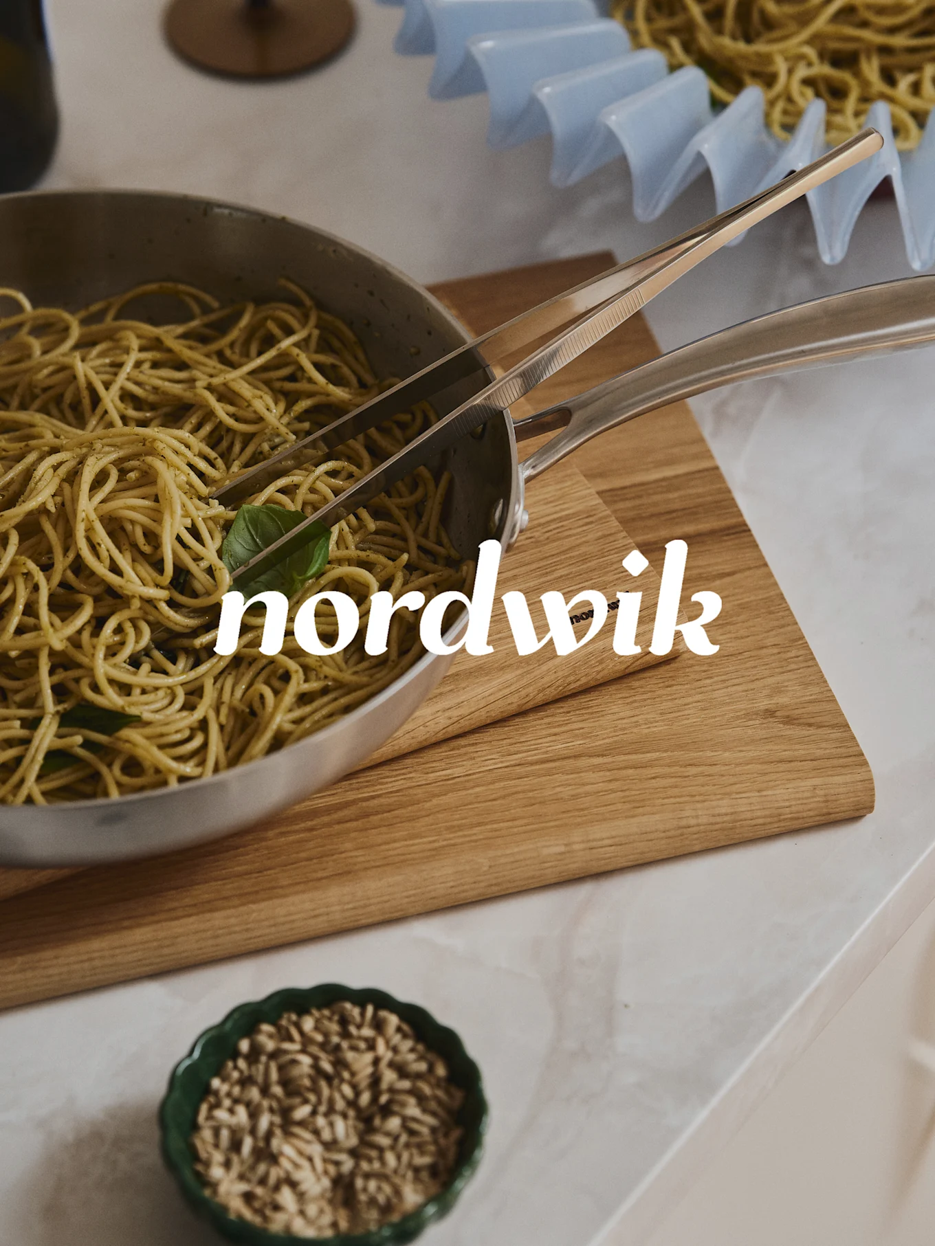 Spaghetti met pesto in een stalen pan met tang op een houten plank. Een kom met zaden eronder, en een pastarek op de achtergrond. 'nordwik' tekstoverlay.