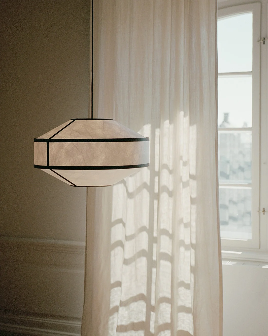 Een geometrische papieren hanglamp met donkere lijsten hangt naast een zonovergoten, doorzichtig gordijn en raam.
