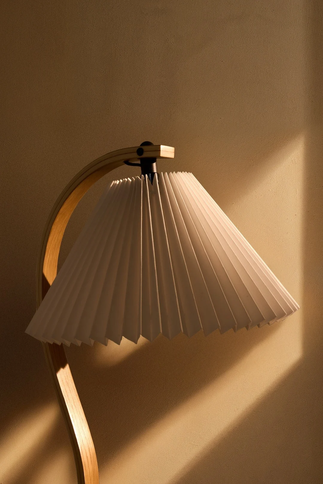 Een lamp met een witte geplooide kap en een gebogen houten voet, die warm licht werpt op een getextureerde muur.