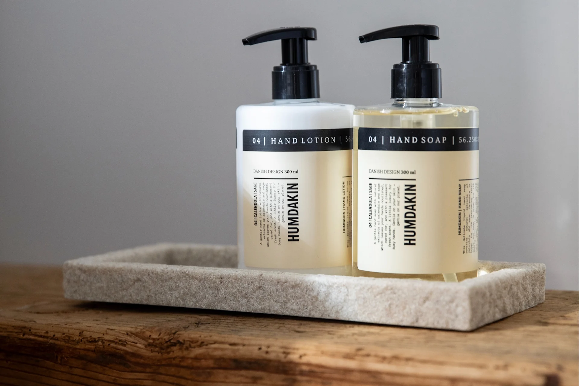 Twee Humdakin pompflessen, handlotion (wit) en handzeep (helder), op een steenachtig dienblad op hout.