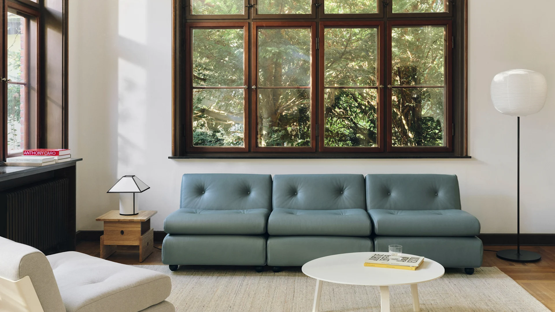 Moderne woonkamer met een blauw-grijze modulaire bank, witte salontafel en een groot raam met uitzicht op groene bomen. Twee lampen verlichten de ruimte.