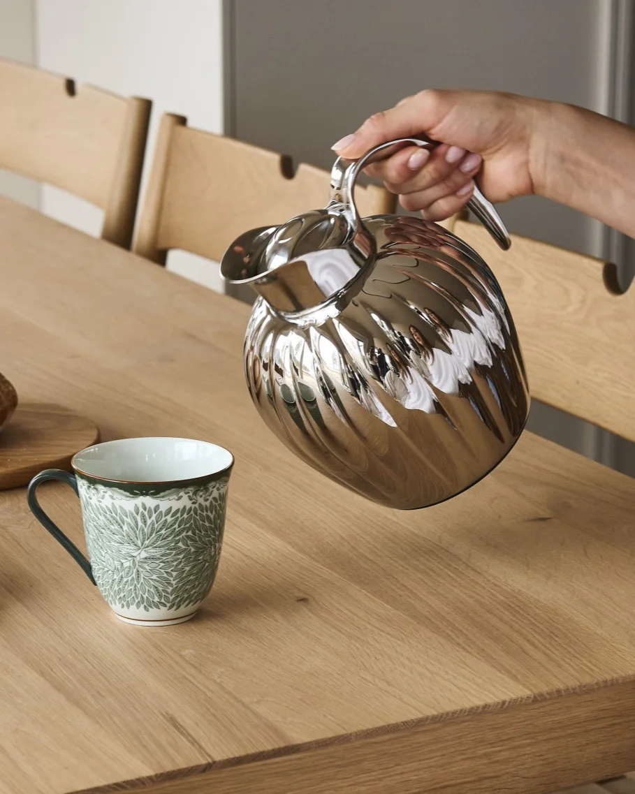 Een hand giet uit een zilveren geribbelde karaf in een witte mok met groen bladmotief op een houten tafel.