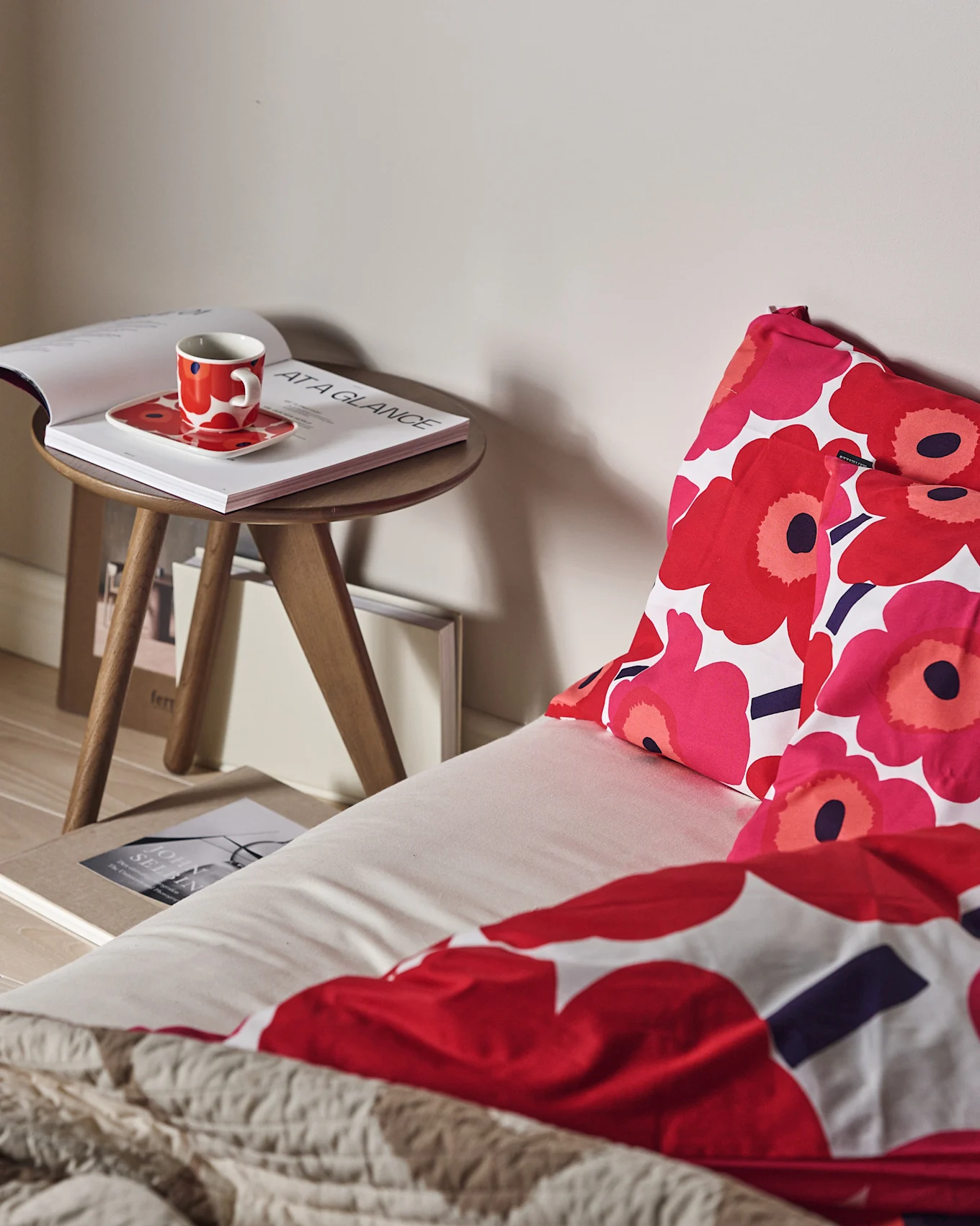 Een bed met rood en roze bloemendekbed en een getextureerde beige plaid. Een rode kop en open boek op een houten bijzettafel.