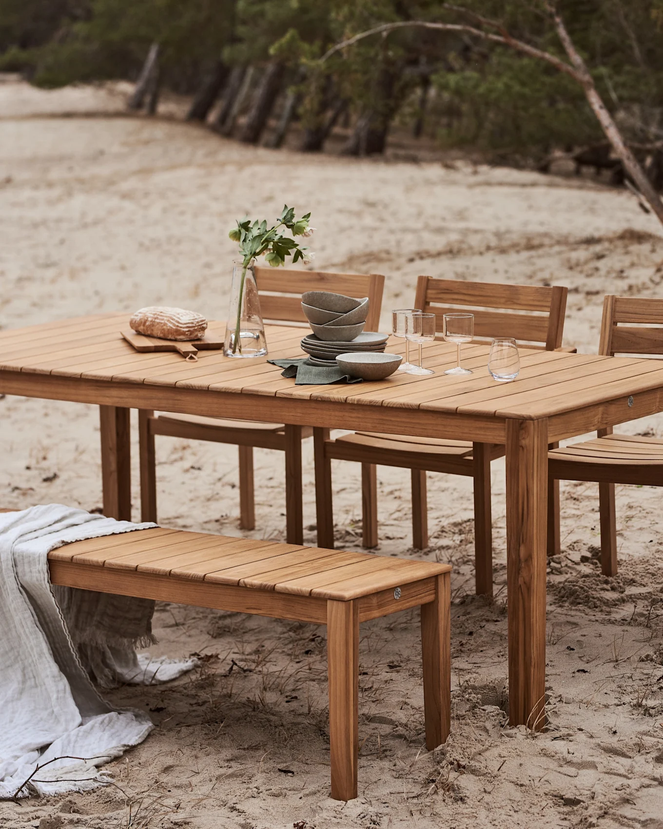 Houten buitenmeubelset met een tafel en bank op een zandstrand. De tafel is gedekt met servies, glazen, brood en een vaas met bloemen.