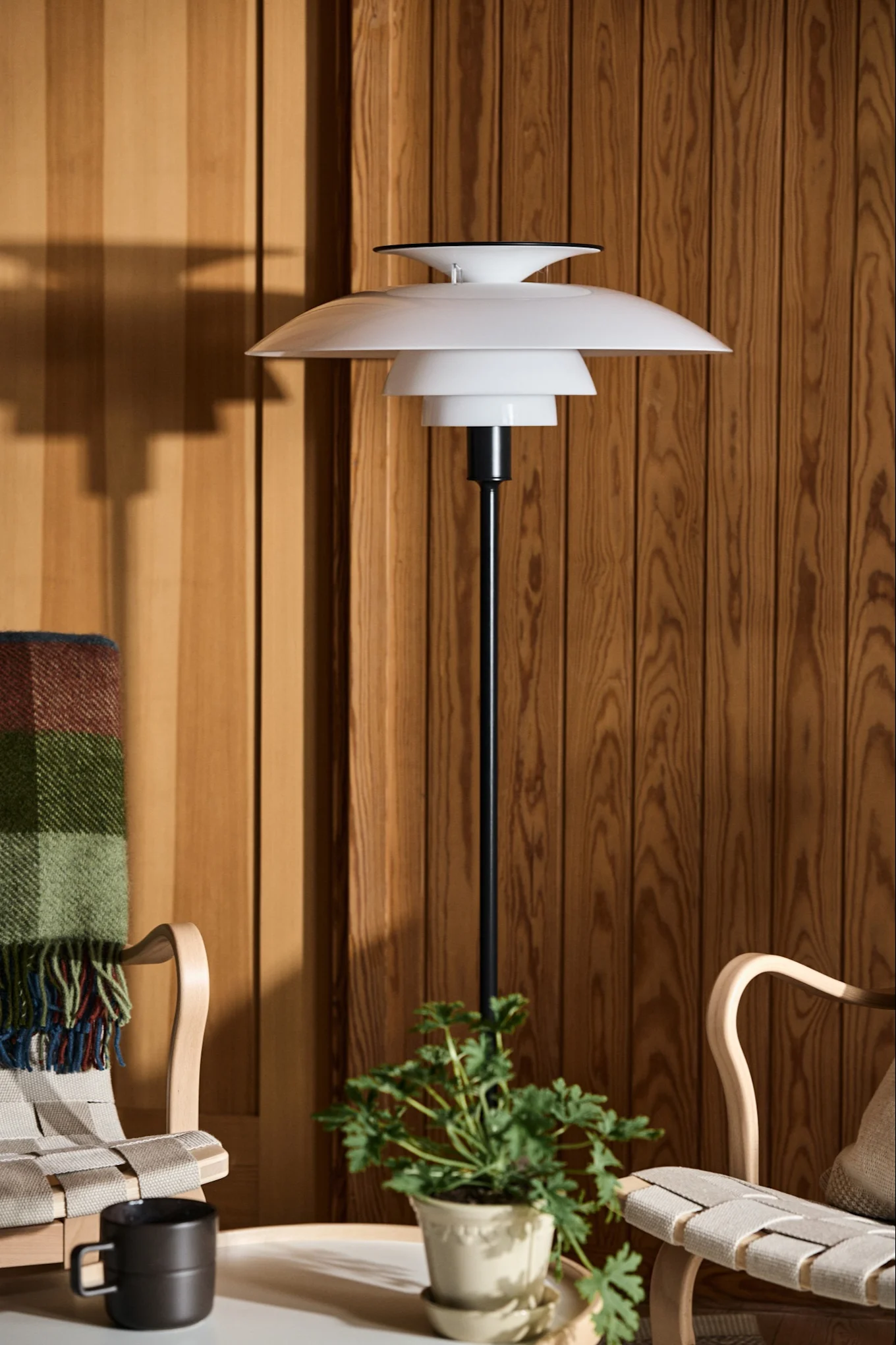 Witte gelaagde vloerlamp naast twee houten stoelen en een plant tegen een houten lambrisering.